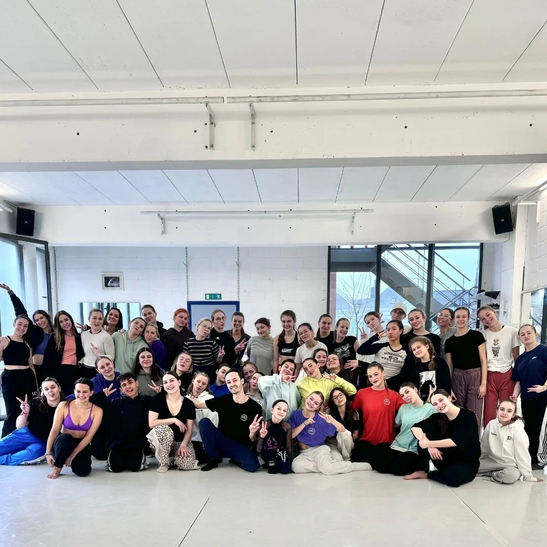 Groupe de jeunes danseurs posant dans une salle de danse lumineuse équipée de grands miroirs et fenêtres, certains faisant des signes de paix ou des gestes joyeux.