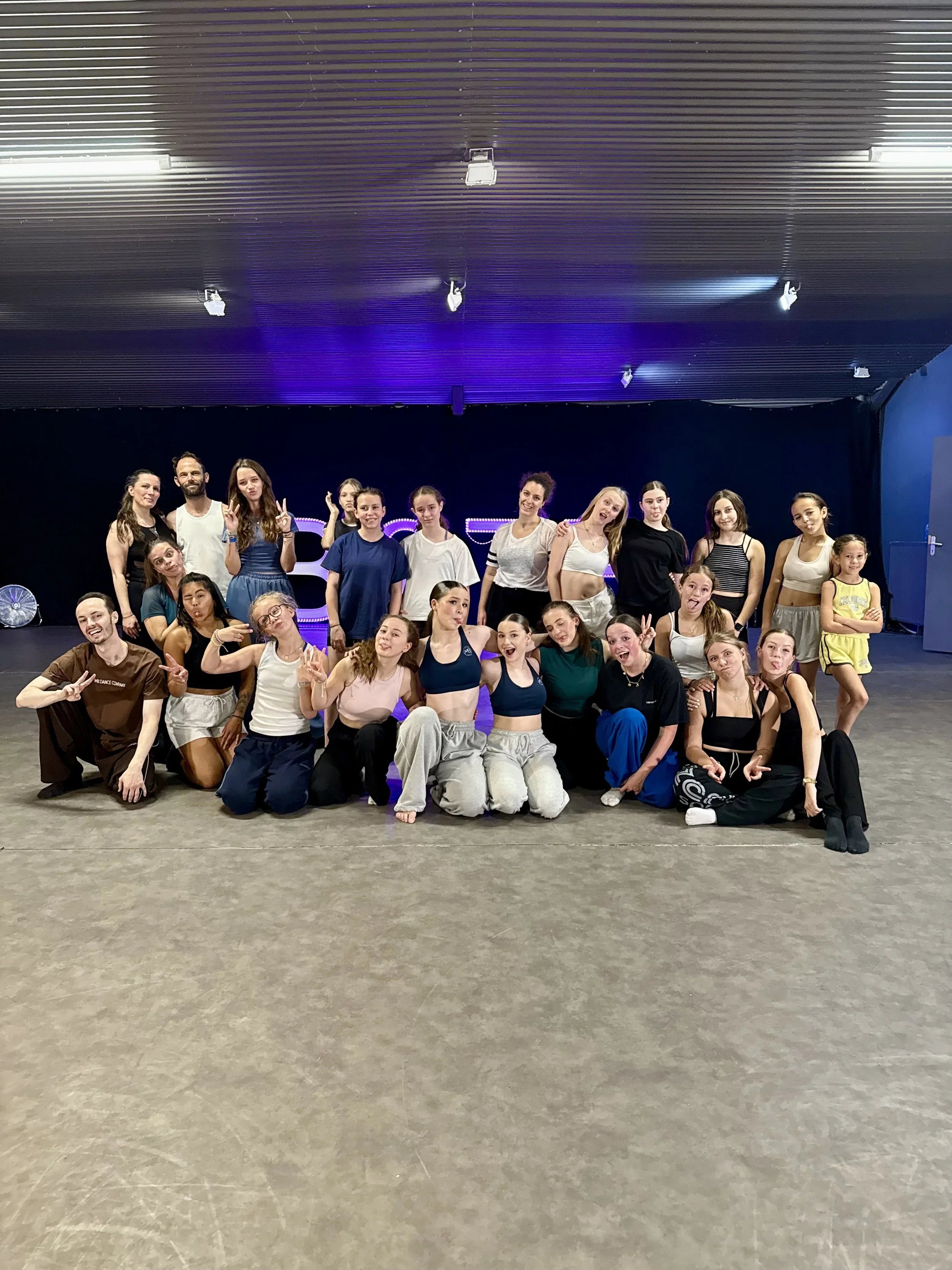 Groupe de jeunes danseurs et danseuses posant pour une photo dans une salle de danse, certains faisant des poses amusantes, derrière eux un fond avec un logo lumineux.
