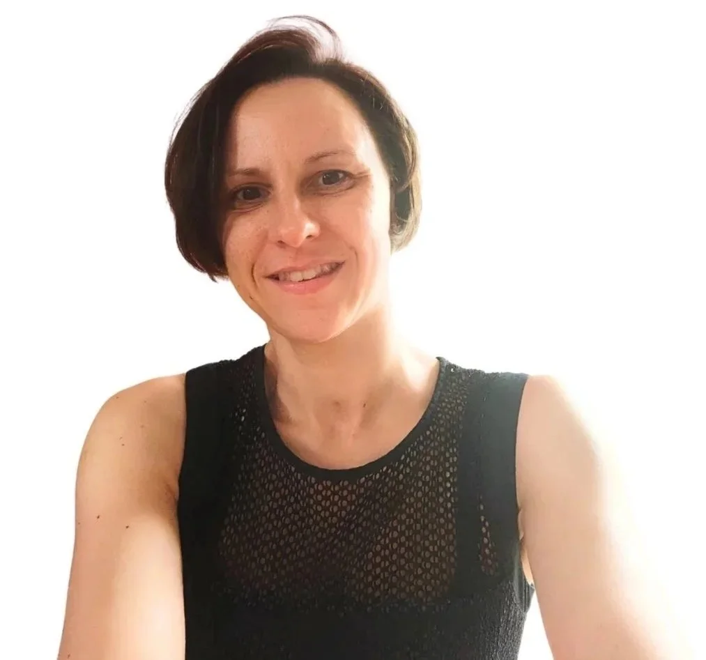 Une femme souriante avec des cheveux courts, portant un haut en filet noir, posant pour une photo en intérieur.