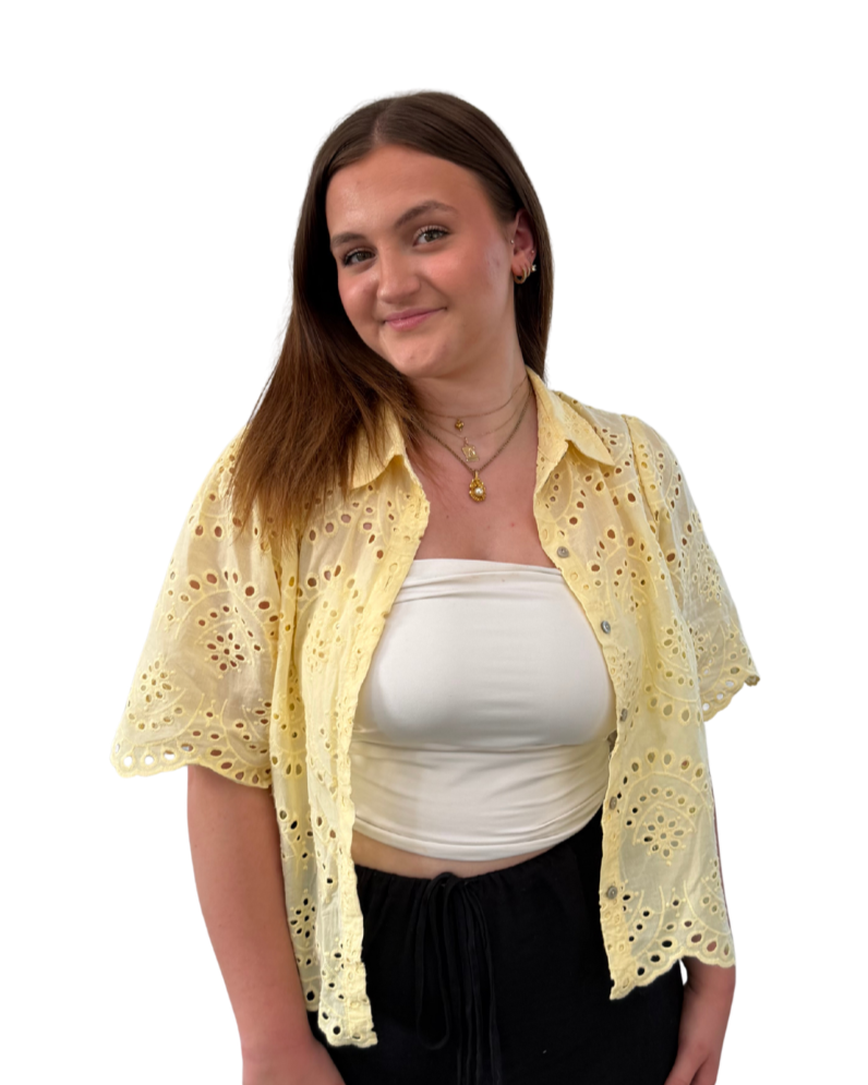 Une jeune femme souriante avec des cheveux bruns, portant un haut blanc, une veste jacquard jaune pâle avec des trous décoratifs et un pantalon noir avec un cordon de serrage.