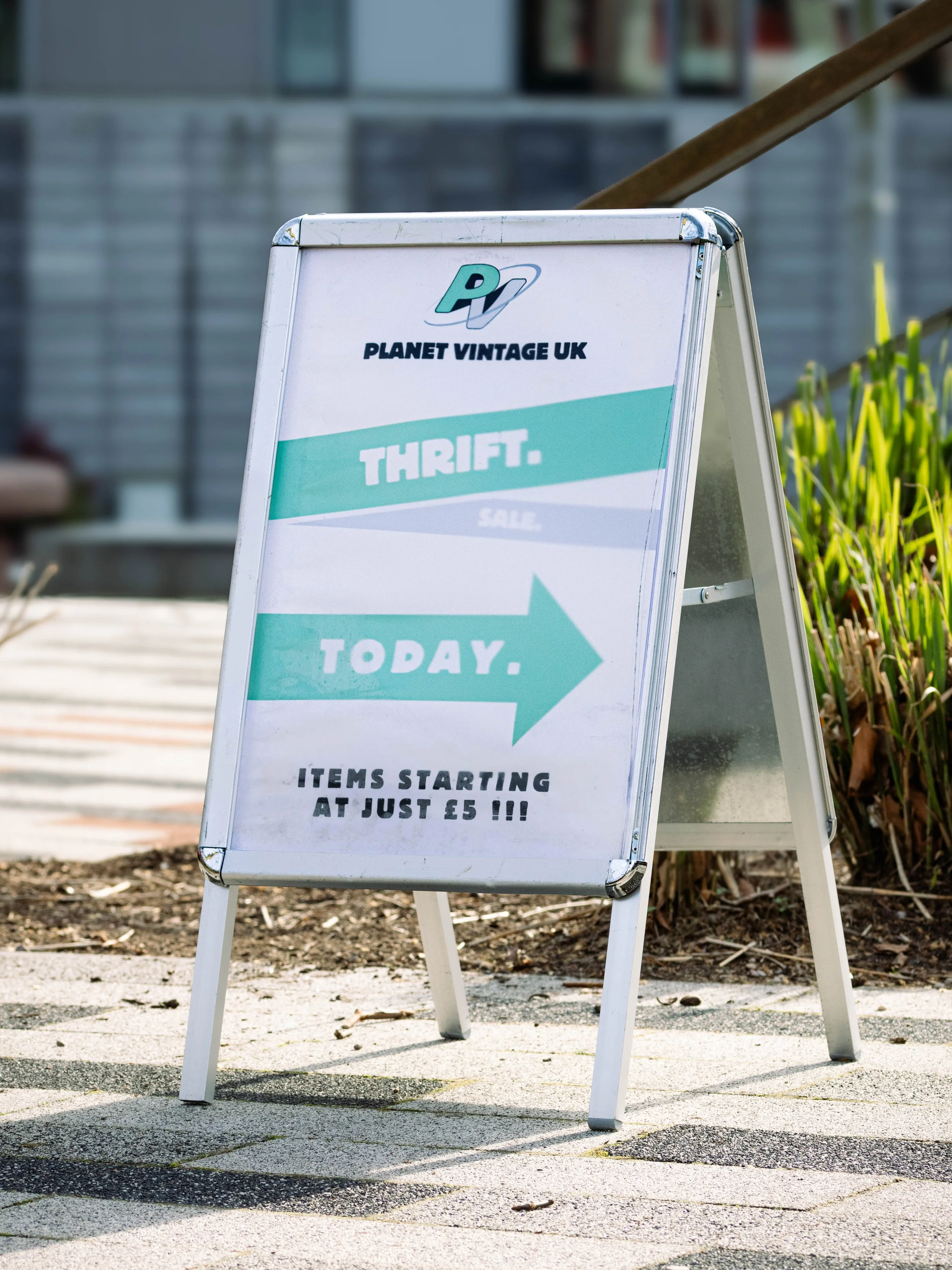 A white sandwich board sign outdoors advertising a thrift store called Planet Vintage UK. The sign promotes thrift shopping with items starting at just five pounds and directs people to the store today. The sign is placed on a paved surface near some