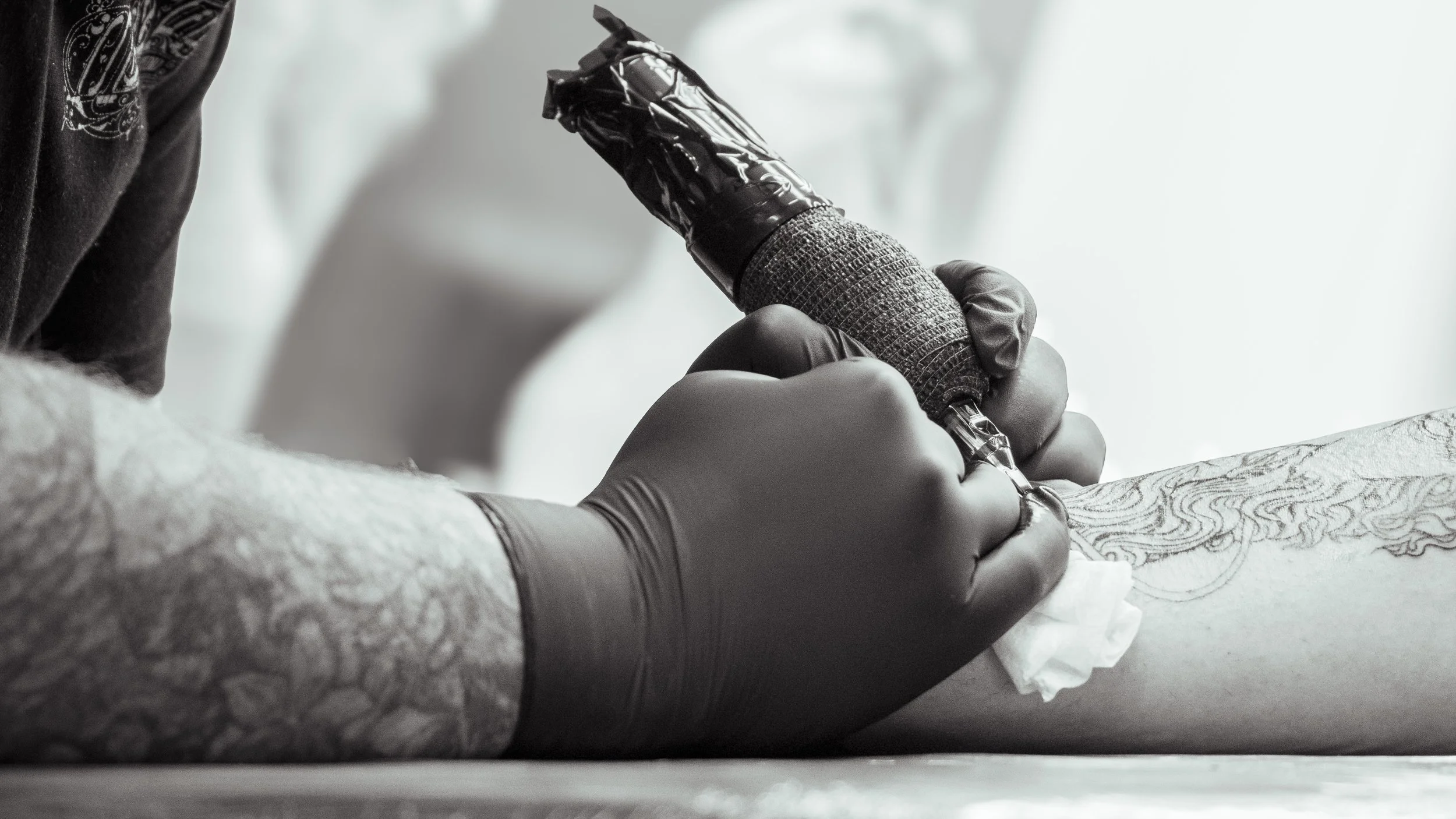 Person with tattoos tattooing another person's arm with a tattoo machine wearing black gloves.