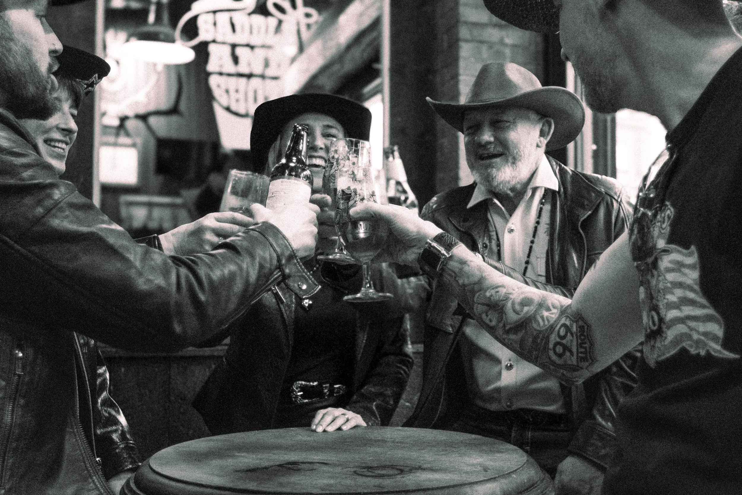 A group of five friends, three men and two women, gathered around a table in a bar, raising their glasses in a toast. They are smiling and appear to be enjoying themselves. The man on the far right is wearing sunglasses and has tattoos on his arm. Th