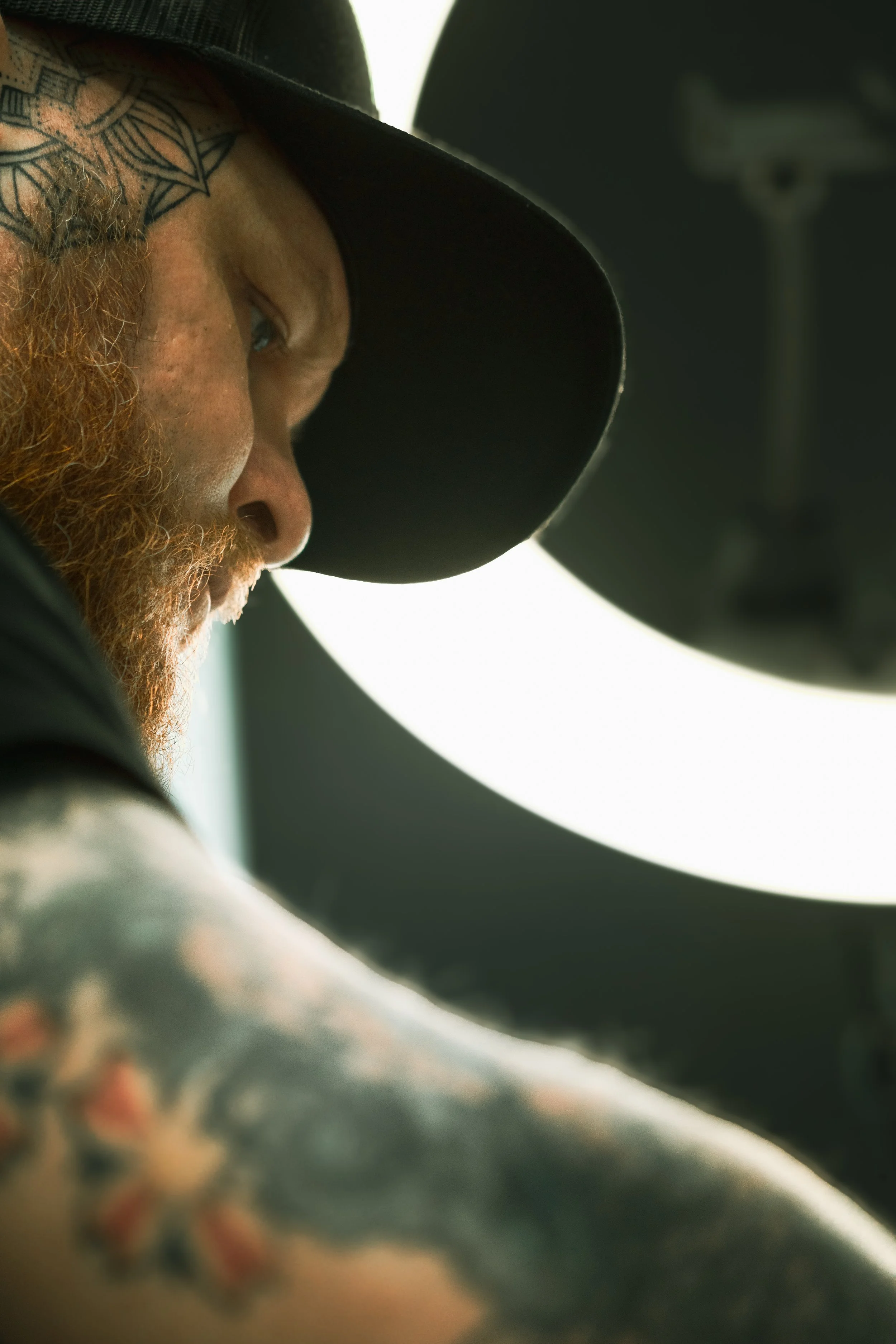 Close-up of a person's face with a tattoo on the forehead, wearing a black hat, with a blurred background.