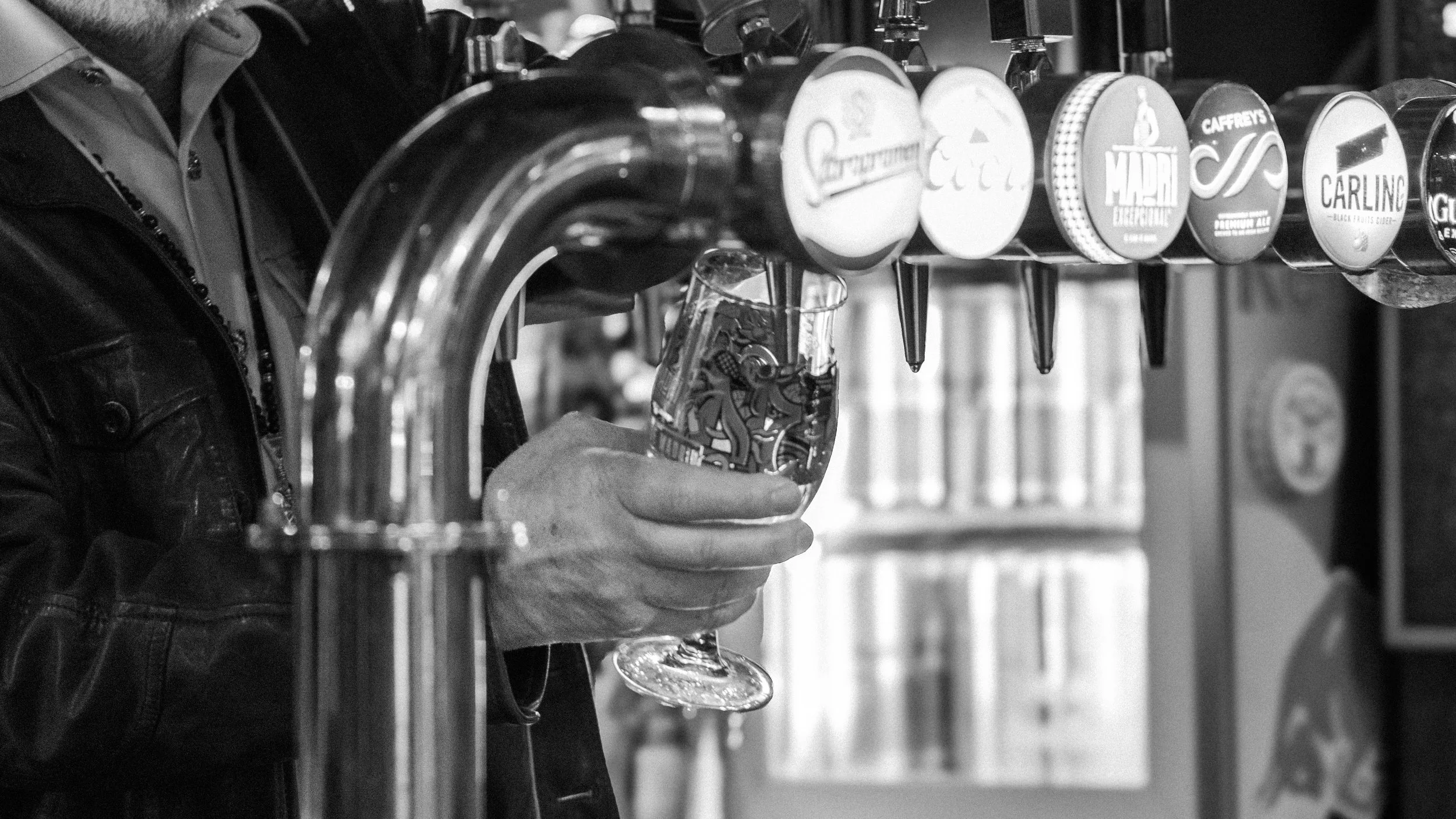 A person holding a glass of beer underneath a row of tap handles in black and white.