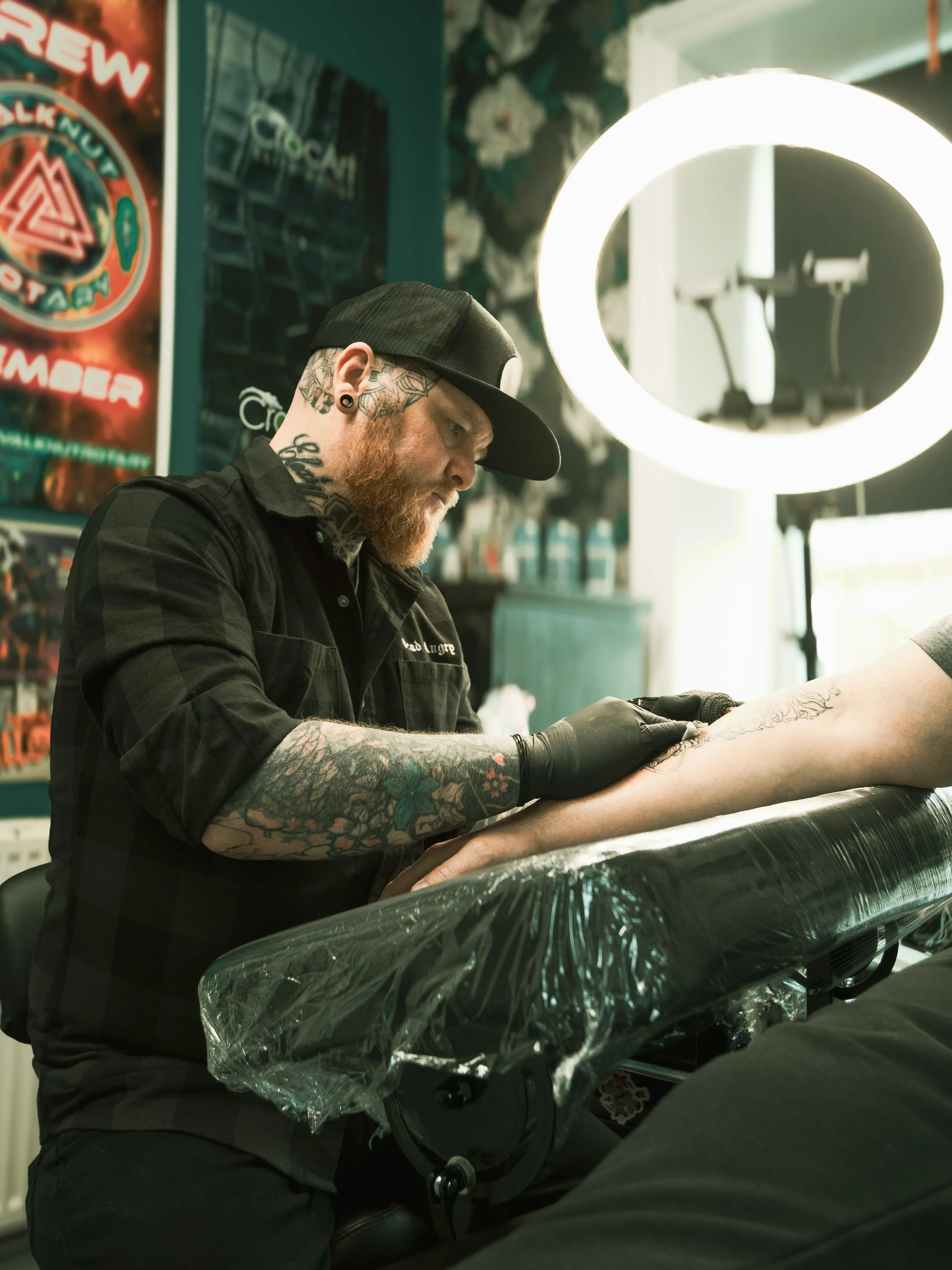 A tattooed man with a beard, wearing a black cap and black shirt, is giving a tattoo to a person's arm at a tattoo parlor illuminated by bright ring light. The background features posters and floral wallpaper.