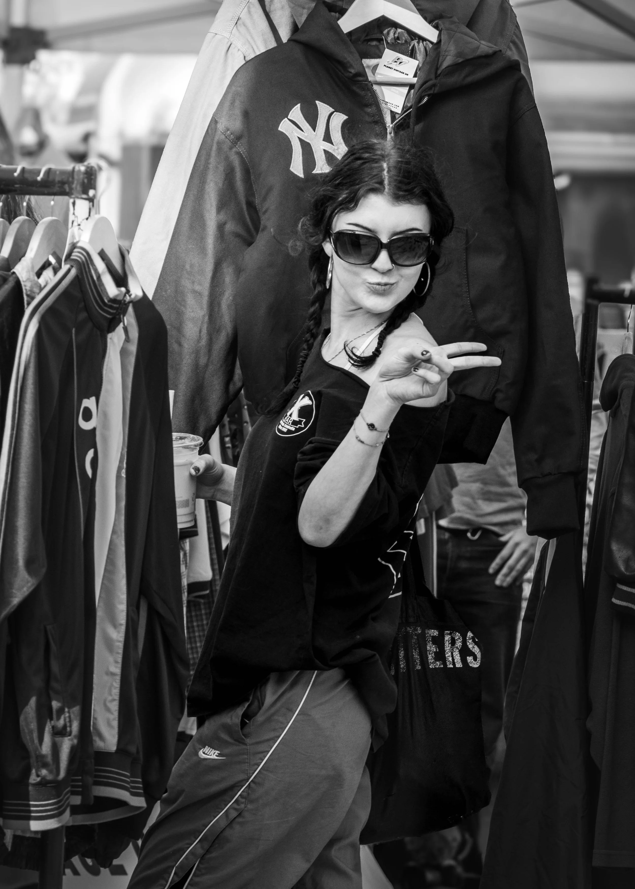 A woman with dark hair and sunglasses making a peace sign at a clothing store, with jackets hanging on racks around her.