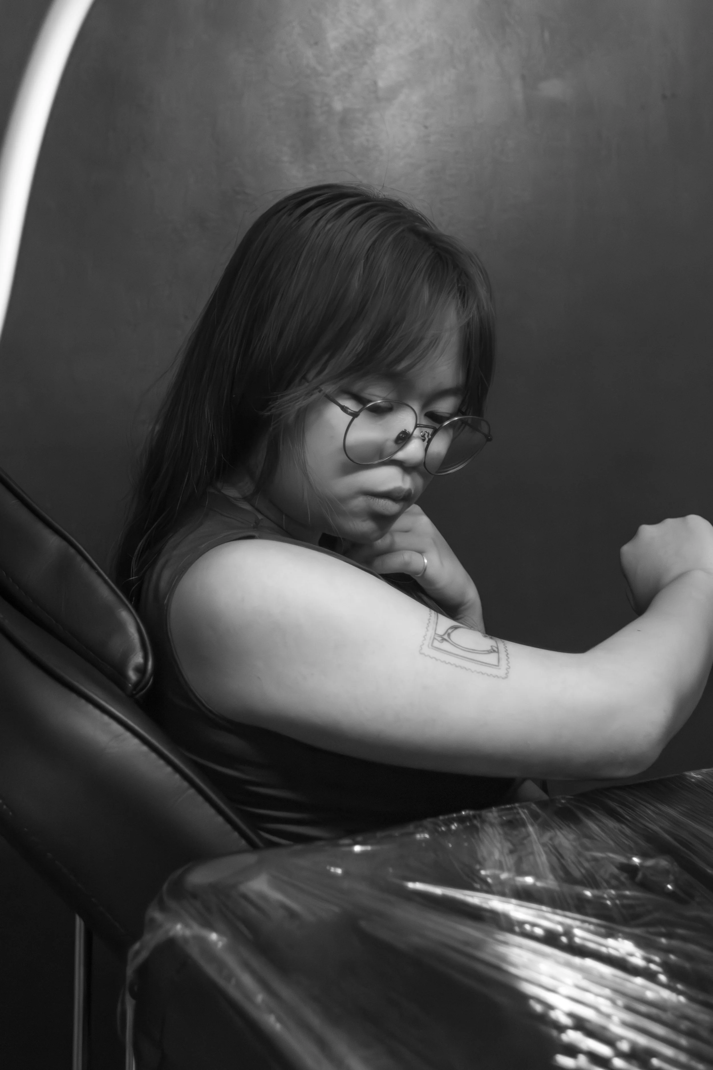 A woman with glasses and dark hair is sitting in a tattoo studio getting a tattoo on her upper arm. She is looking down and appears to be focused, with her hand gently touching her neck. The tattoo is partially visible.