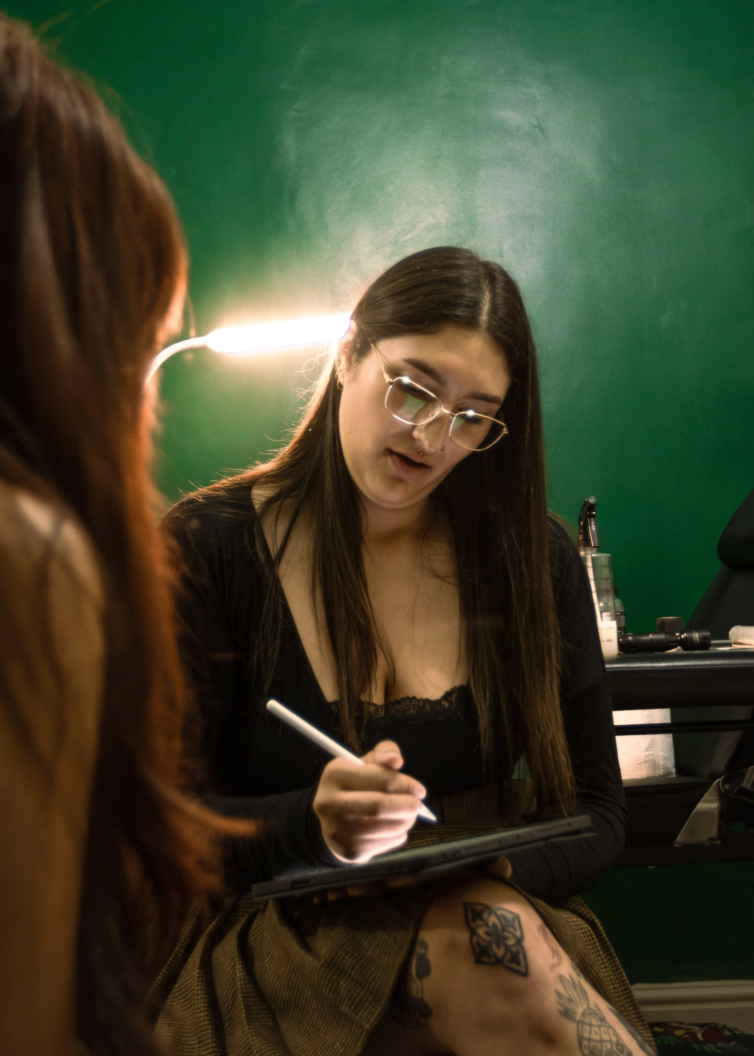 A young woman with long dark hair, glasses, and a septum piercing sitting at a desk, writing on a notepad with a pen, in front of a green wall.