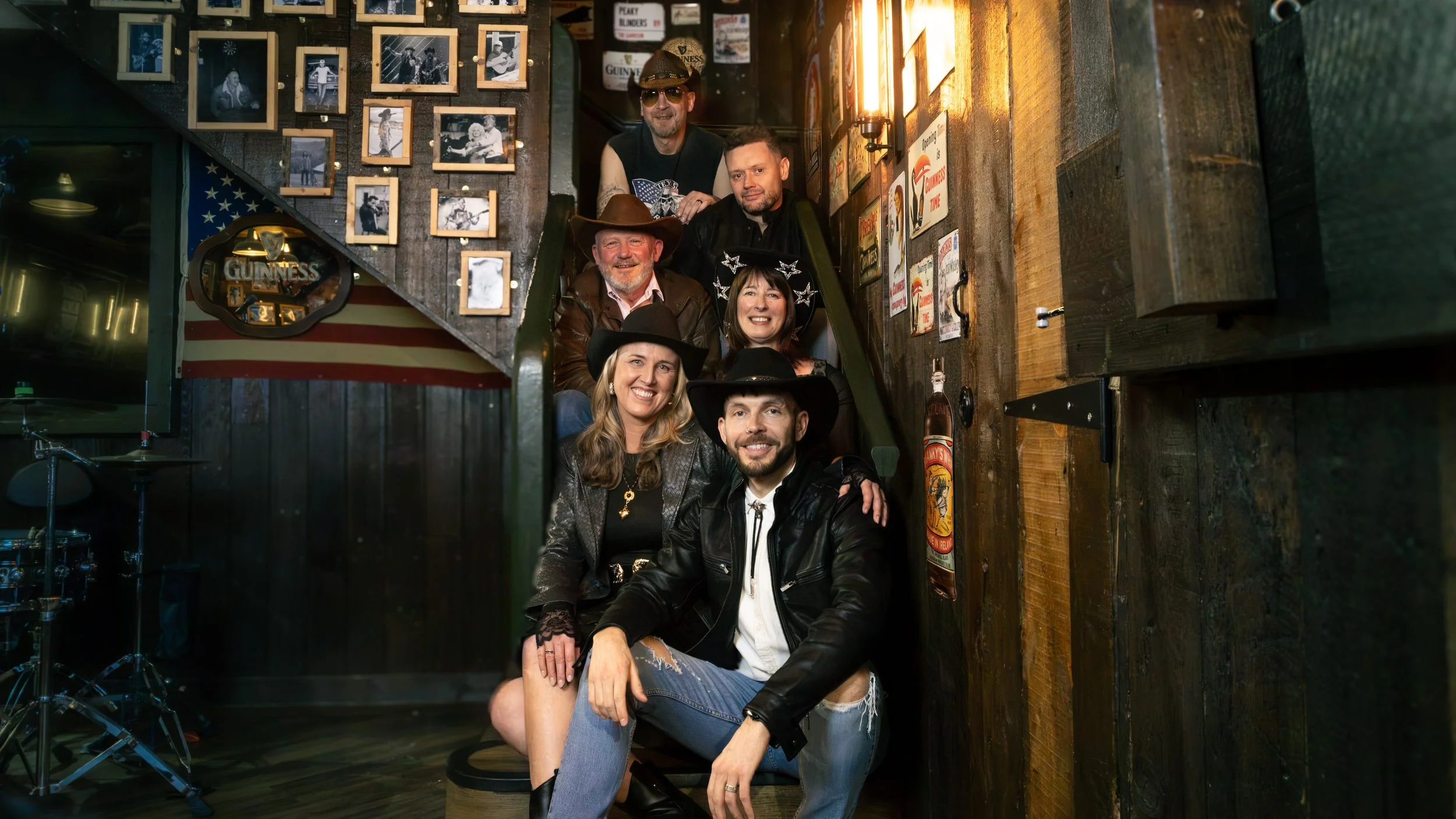 A group of seven people, dressed in casual and country-themed clothing, posing together on a staircase inside a bar or restaurant decorated with photos, signs, and an American flag.