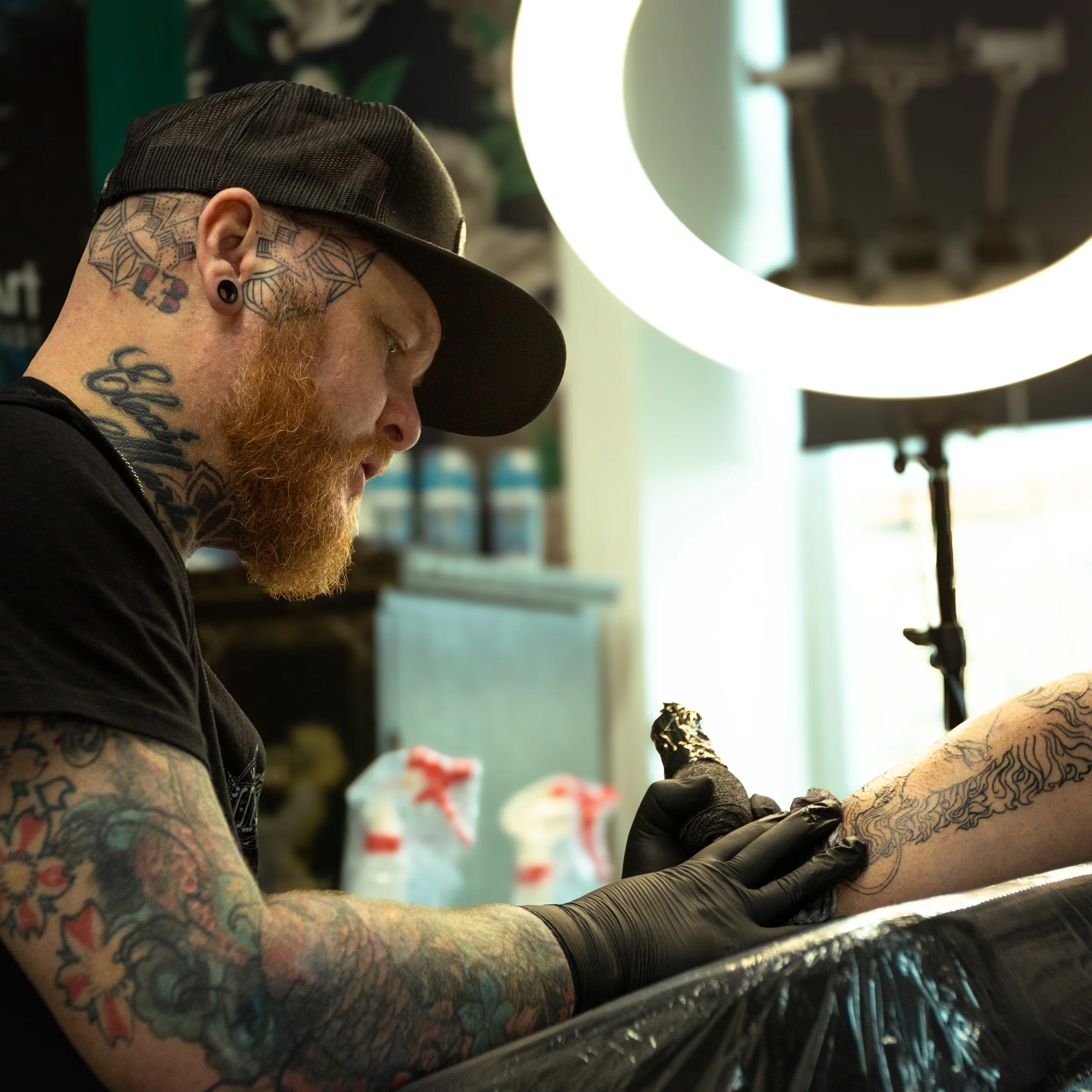 A tattooed person with a beard wearing a black cap and black gloves tattoos another person’s arm.