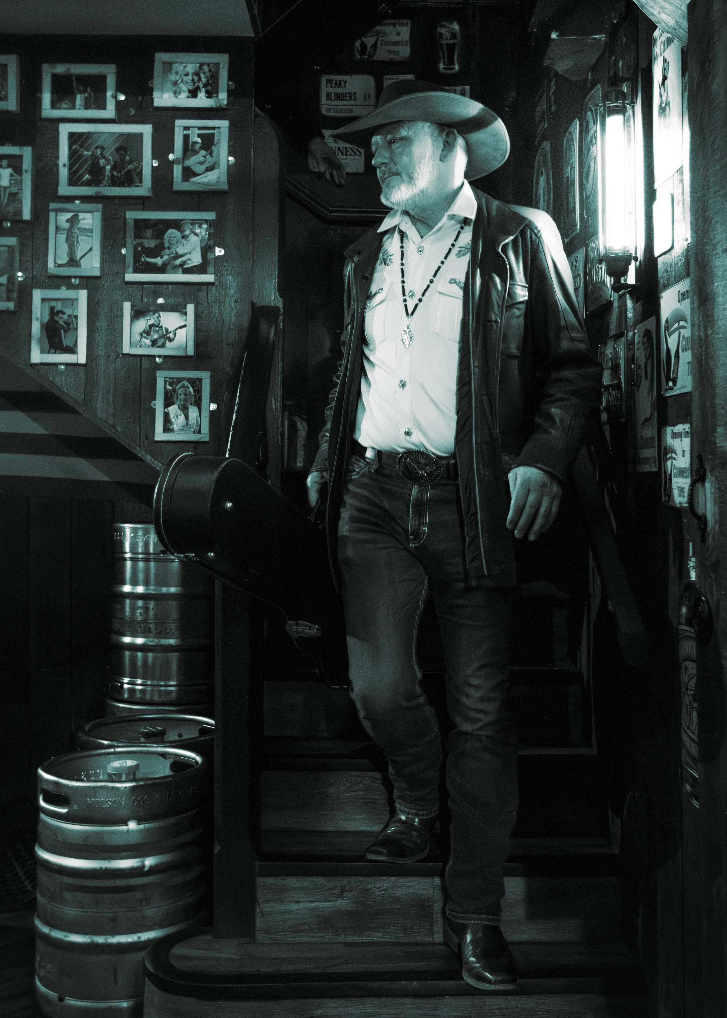 A man with a gray beard wearing a cowboy hat, leather jacket, and beaded necklaces, standing inside a bar or pub with framed photos on the wall and beer kegs on the floor.