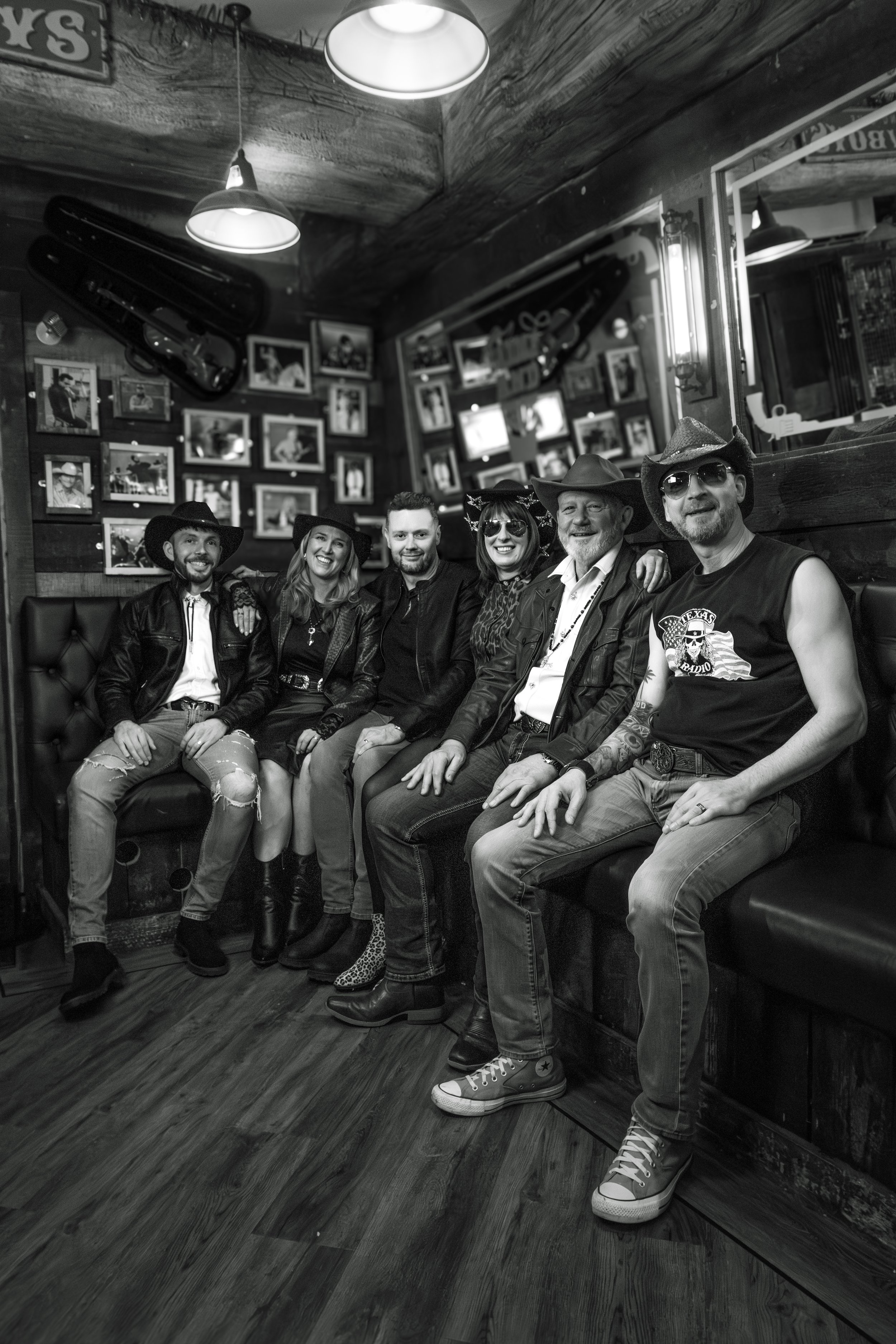 A group of six people sitting on a bench in a bar or restaurant, wearing cowboy hats and casual clothing, smiling for the camera. The wall behind them has framed photographs, and the ceiling has hanging lights.