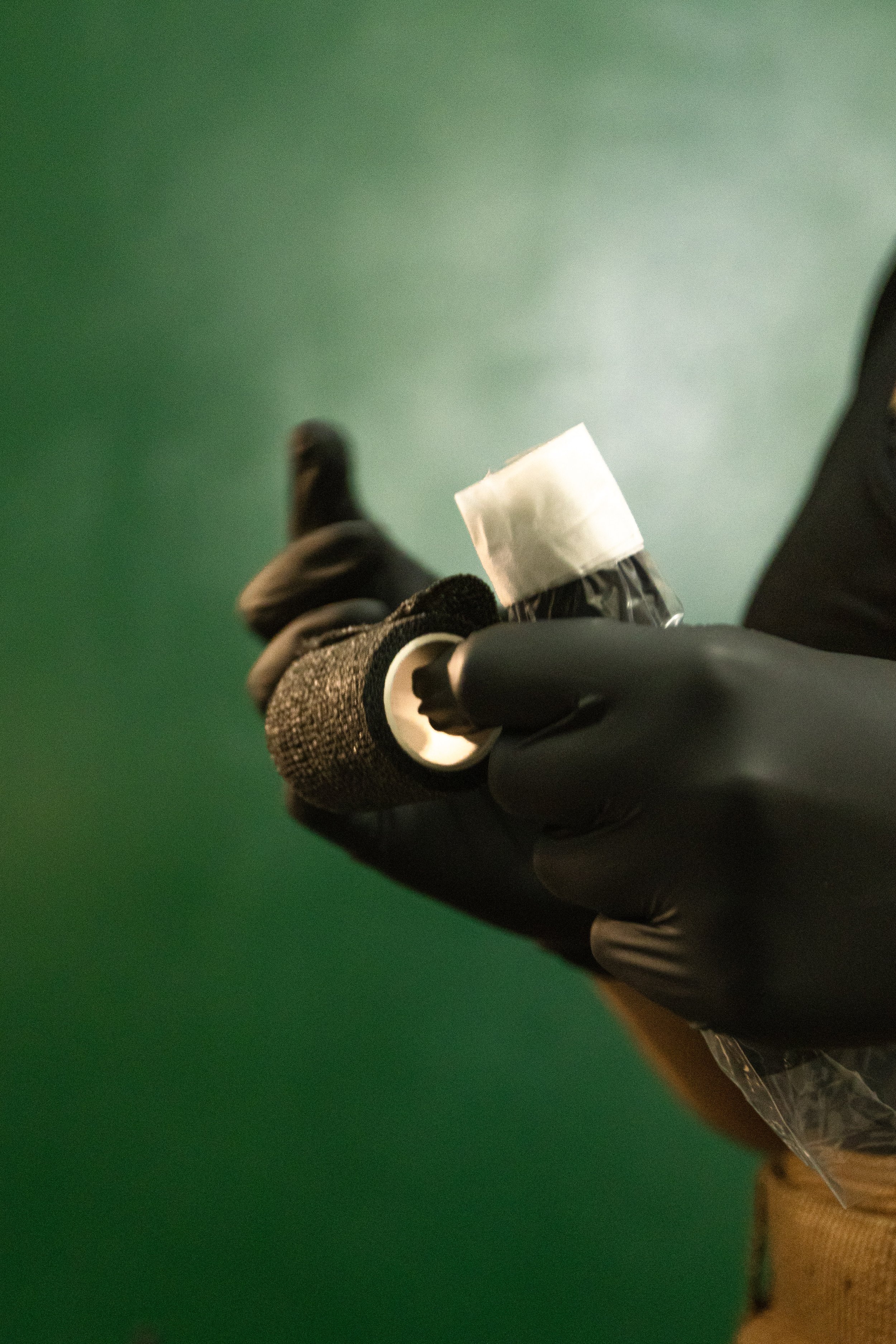 A person wearing a black glove and a brown sweater is holding a small wrapped object with white tape on top over a green background.