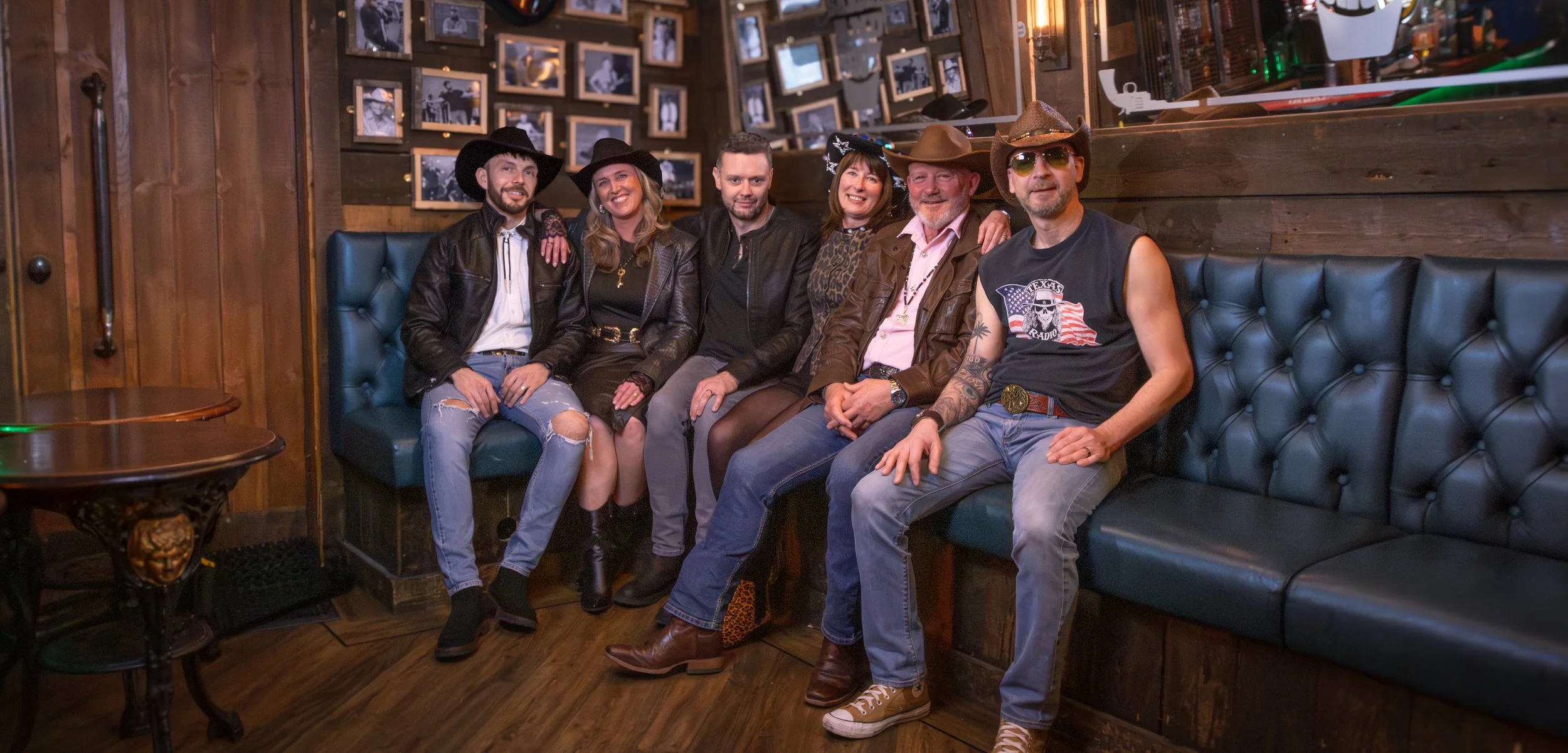 Six people sitting on a teal leather booth in a wooden-paneled bar, dressed in cowboy and western-themed attire, smiling and posing for the photo.