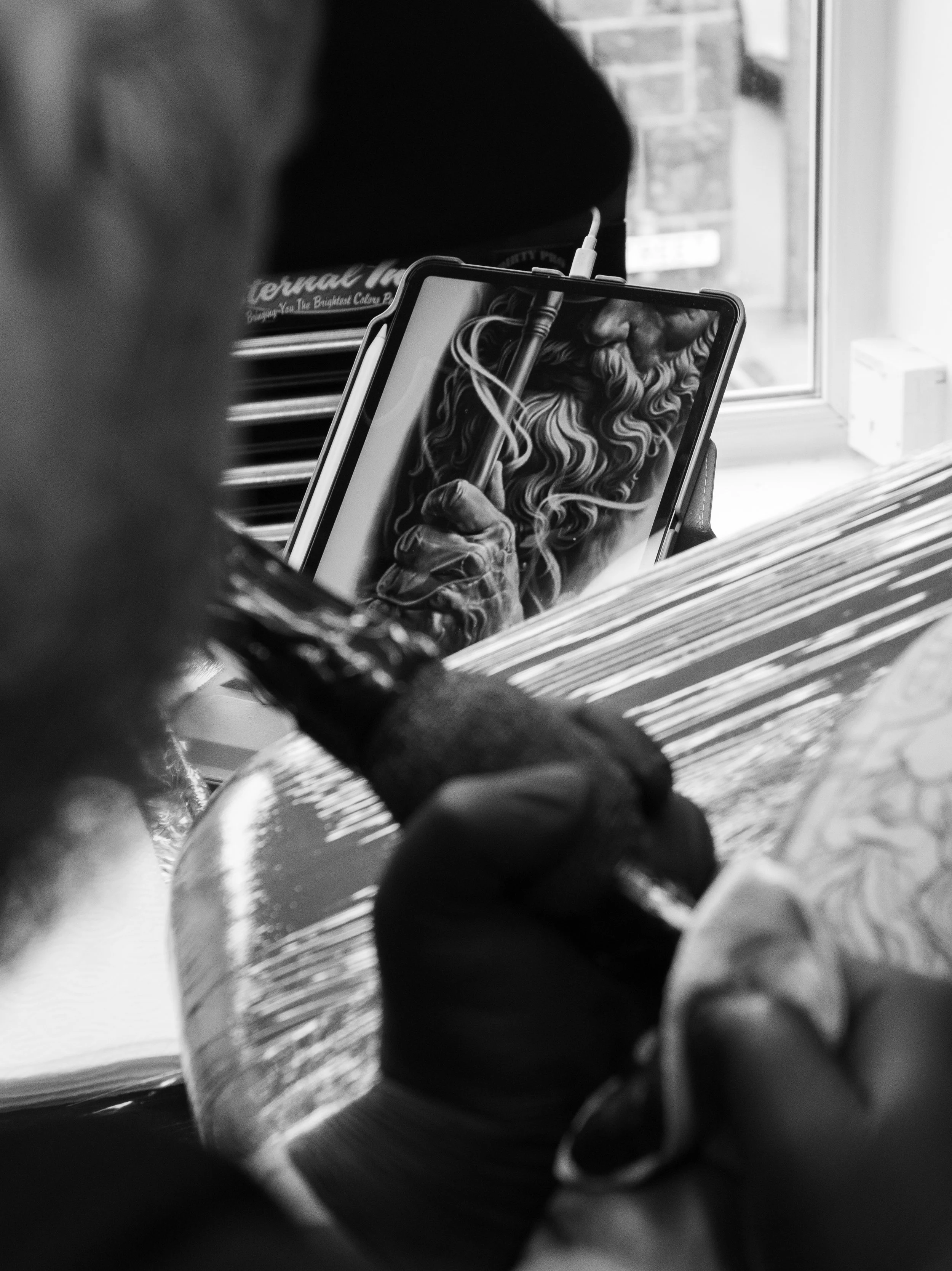 Black and white photo of a smartphone held in a person's hand, displaying a detailed drawing of an old man with a beard, holding a stylus. The background includes a window and part of a brick wall.