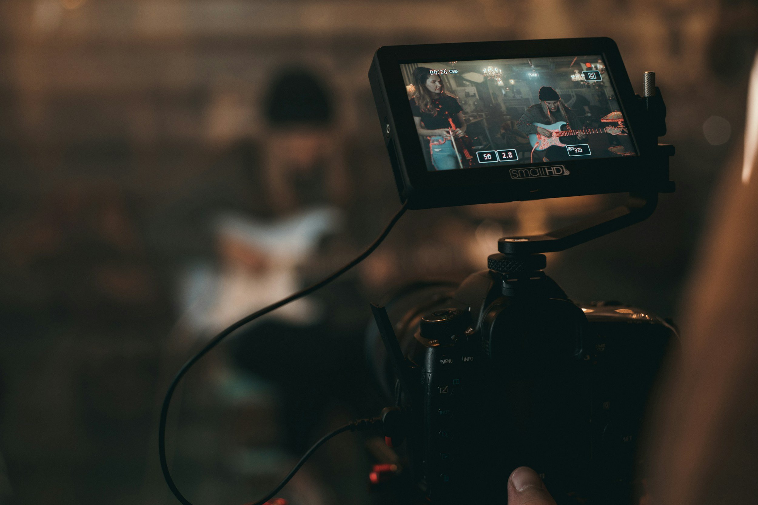 Close-up of a camera monitor showing two women, one playing the guitar and the other holding a phone, in a dimly lit indoor setting.