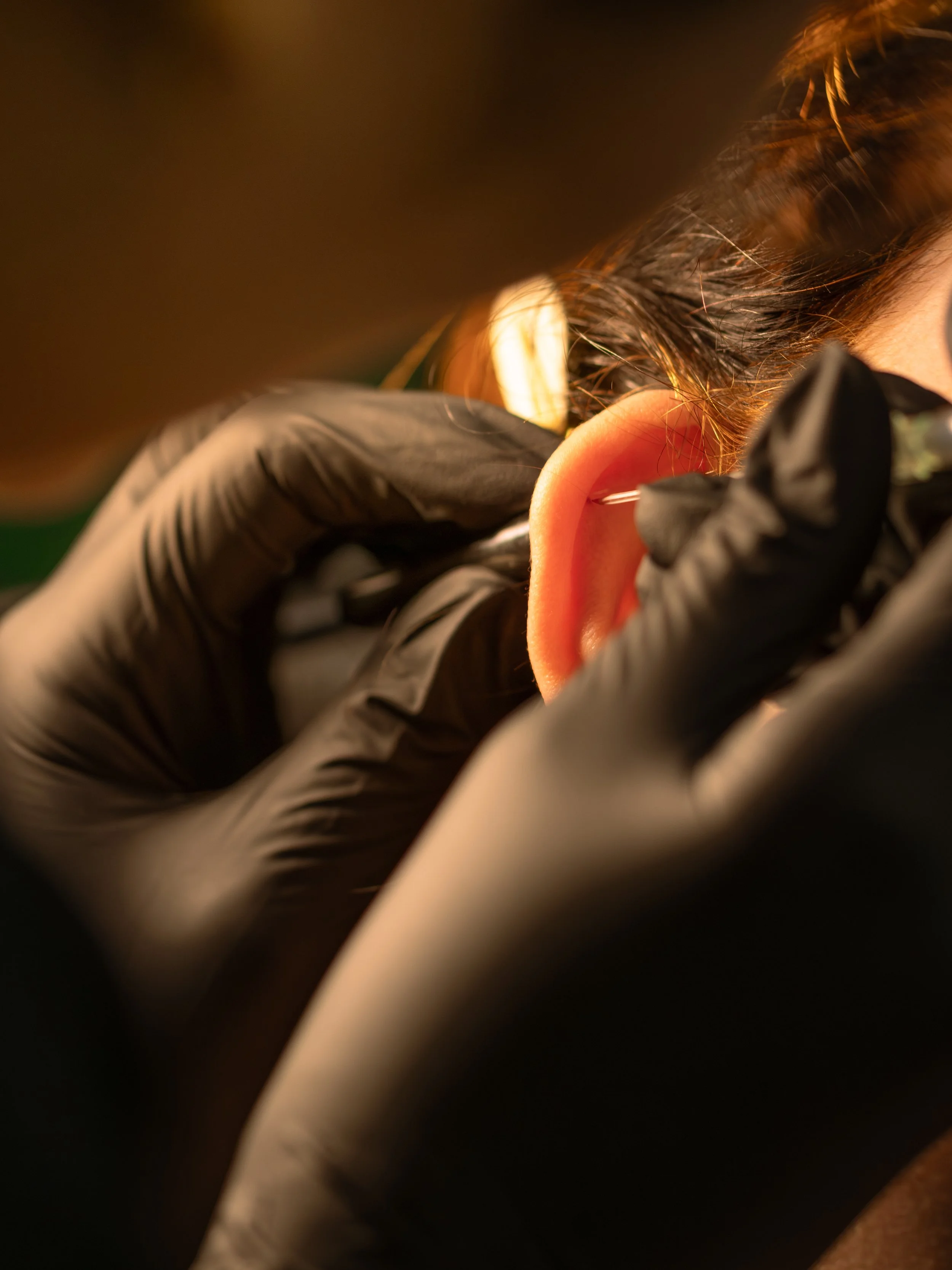 Close-up of a person's ear with a piercings needle being inserted, with someone wearing black gloves performing an ear piercing in a dimly lit environment.