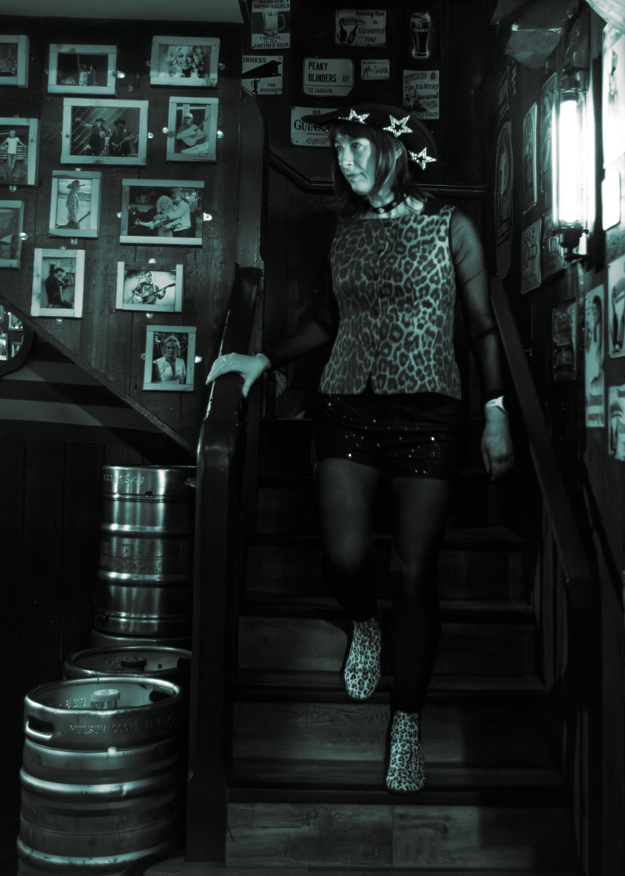 A woman dressed in leopard print clothing, wearing a witch hat with star decorations and leopard print thigh-high boots, standing inside a bar with photos on the wall and beer signs.