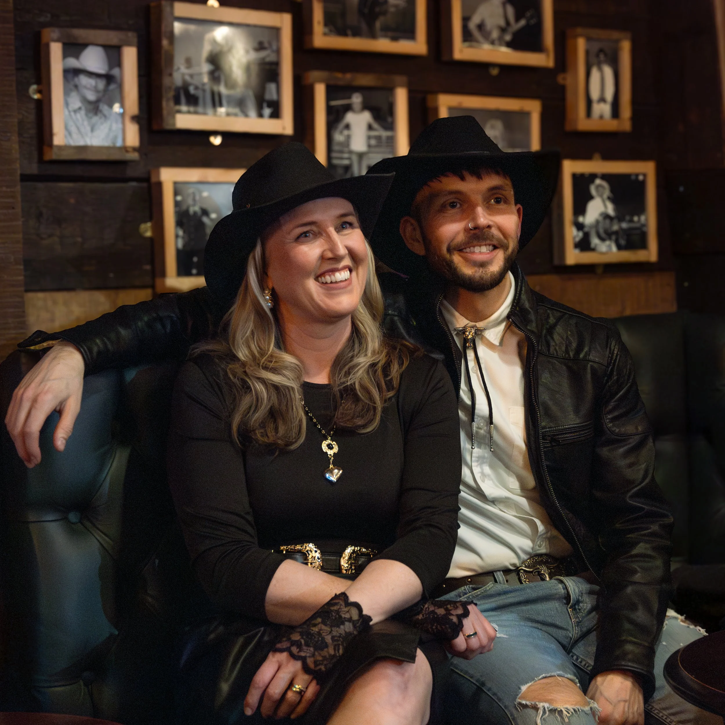 Two friends, a woman and a man, wearing black hats, sitting closely together and smiling in a dimly lit room with a wall decorated with black-and-white photographs in wooden frames.