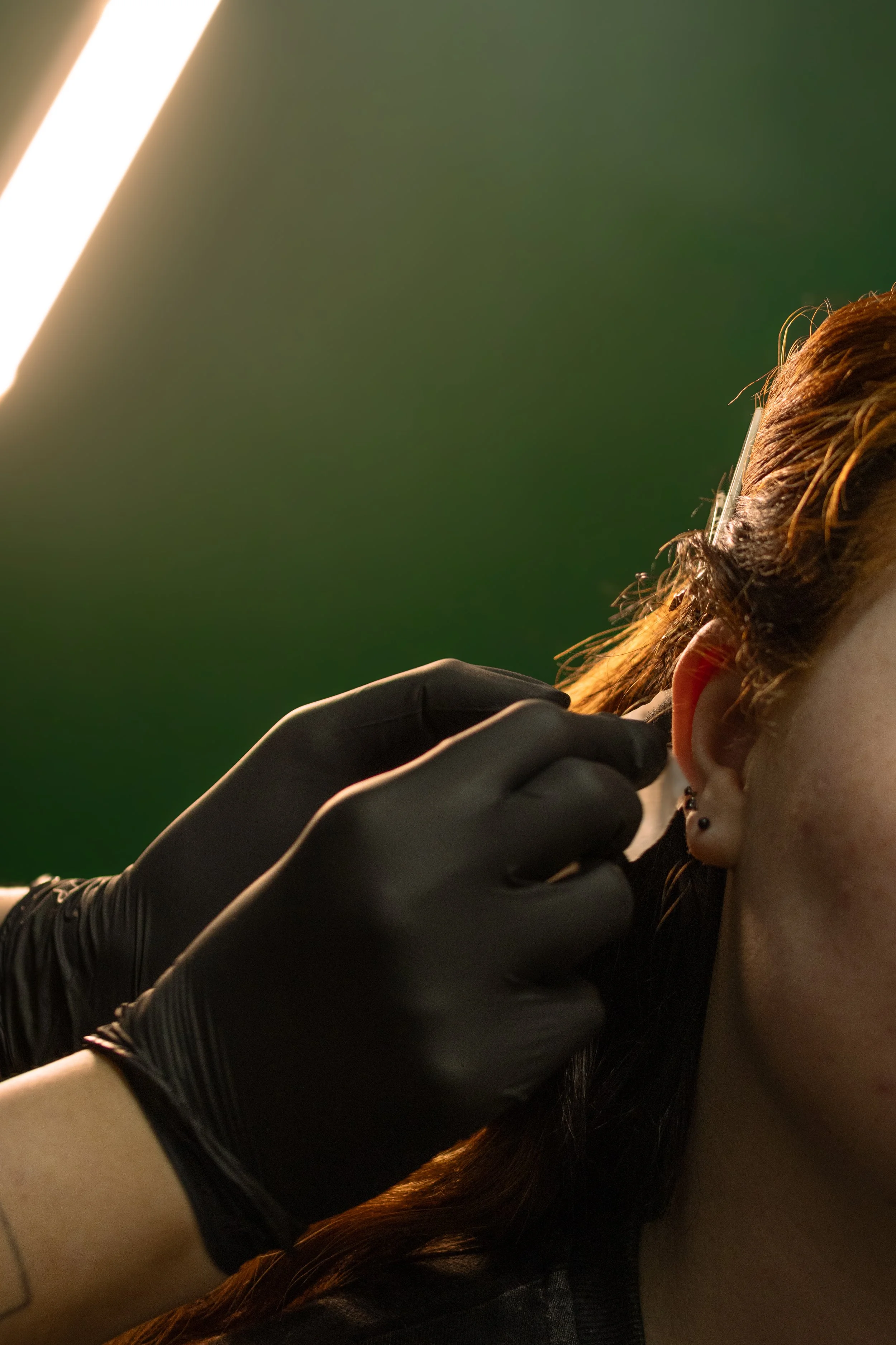 A close-up of a person with long hair and earrings, receiving a tattoo on her neck from a tattoo artist wearing black gloves.