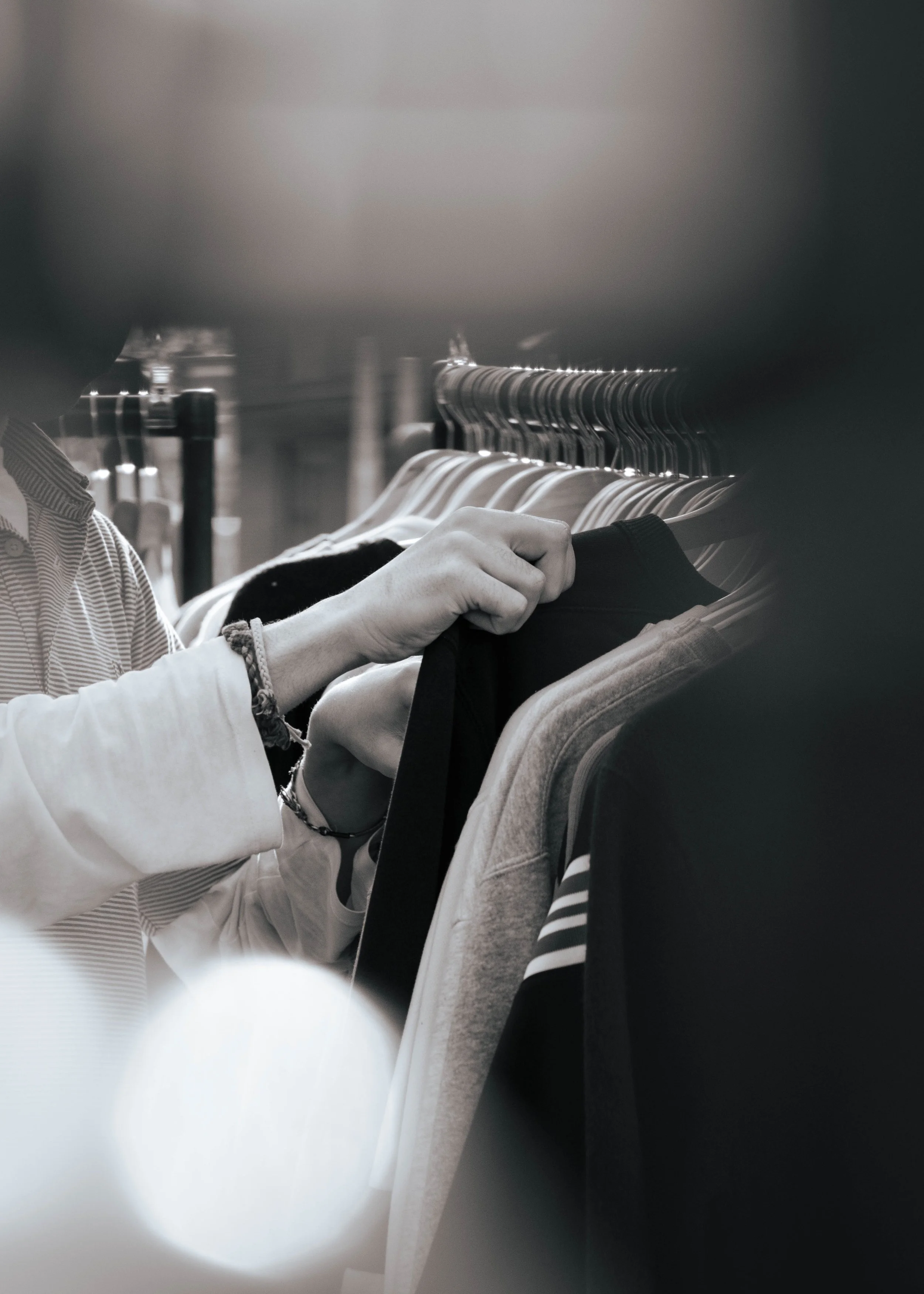 Close-up of a person shopping for clothes, looking at a black shirt on a rack with multiple hangers, in what appears to be a clothing store, with a focus on their hand and part of their arm.