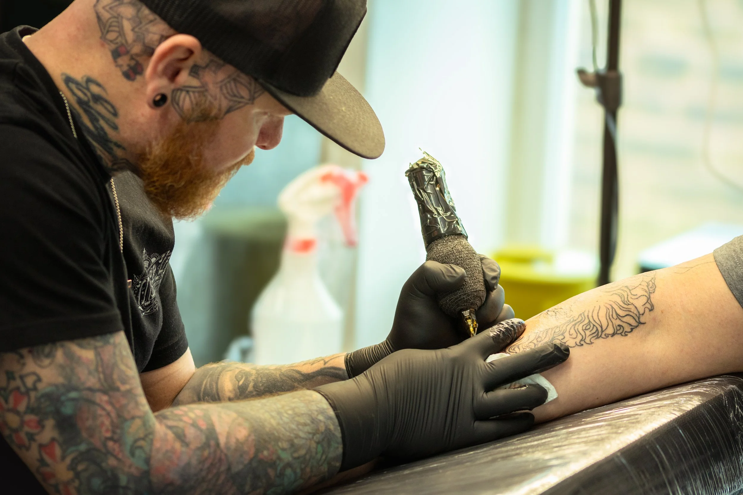 A man with tattoos on his neck and arms, wearing a baseball cap and black gloves, is getting a tattoo on his thigh by a tattoo artist.
