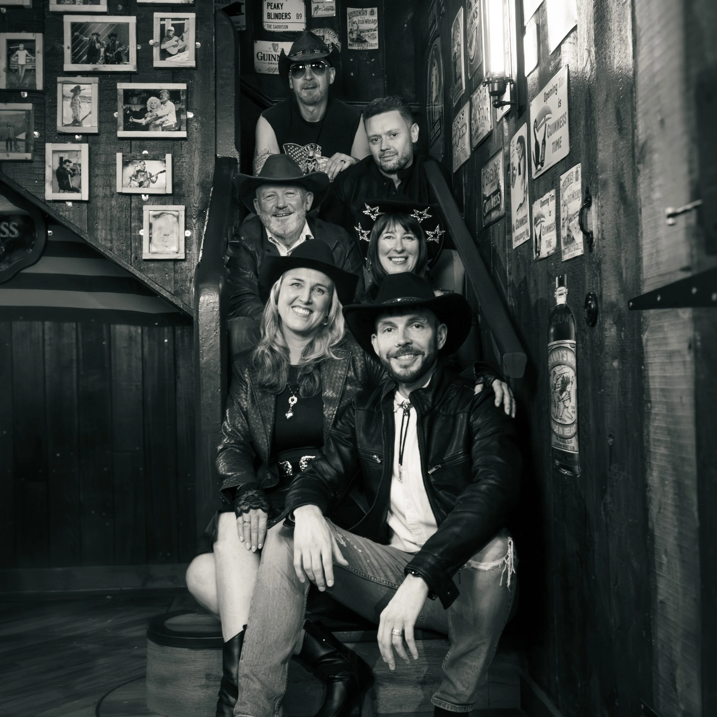 A group of people on a staircase in a bar or pub, wearing casual clothing and hats, with a wall decorated with framed photos and vintage signs in the background.