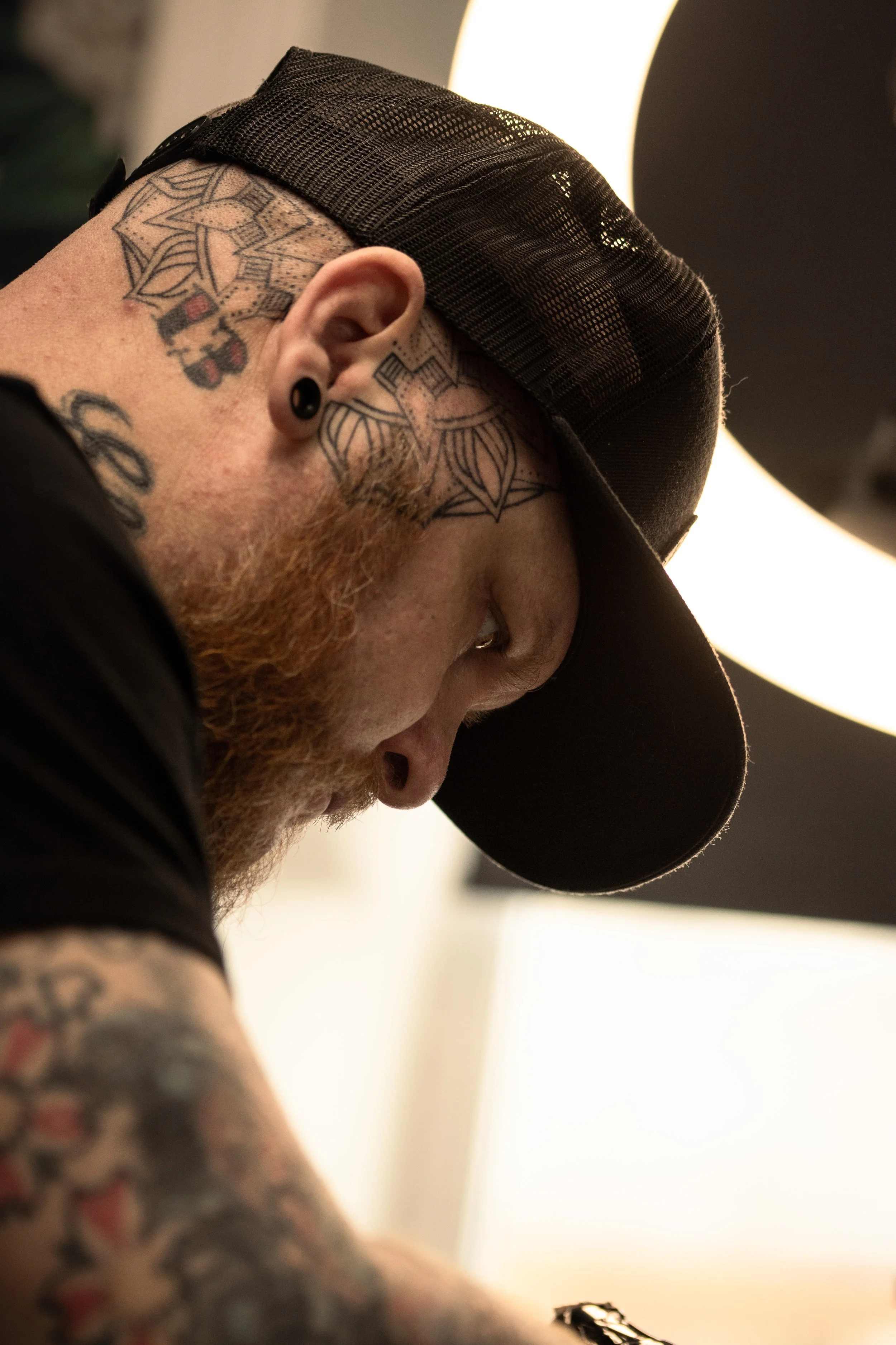 Close-up of a person with tattoos on their neck and arm, wearing a black cap and black earrings, with a blurred background and bright lighting.
