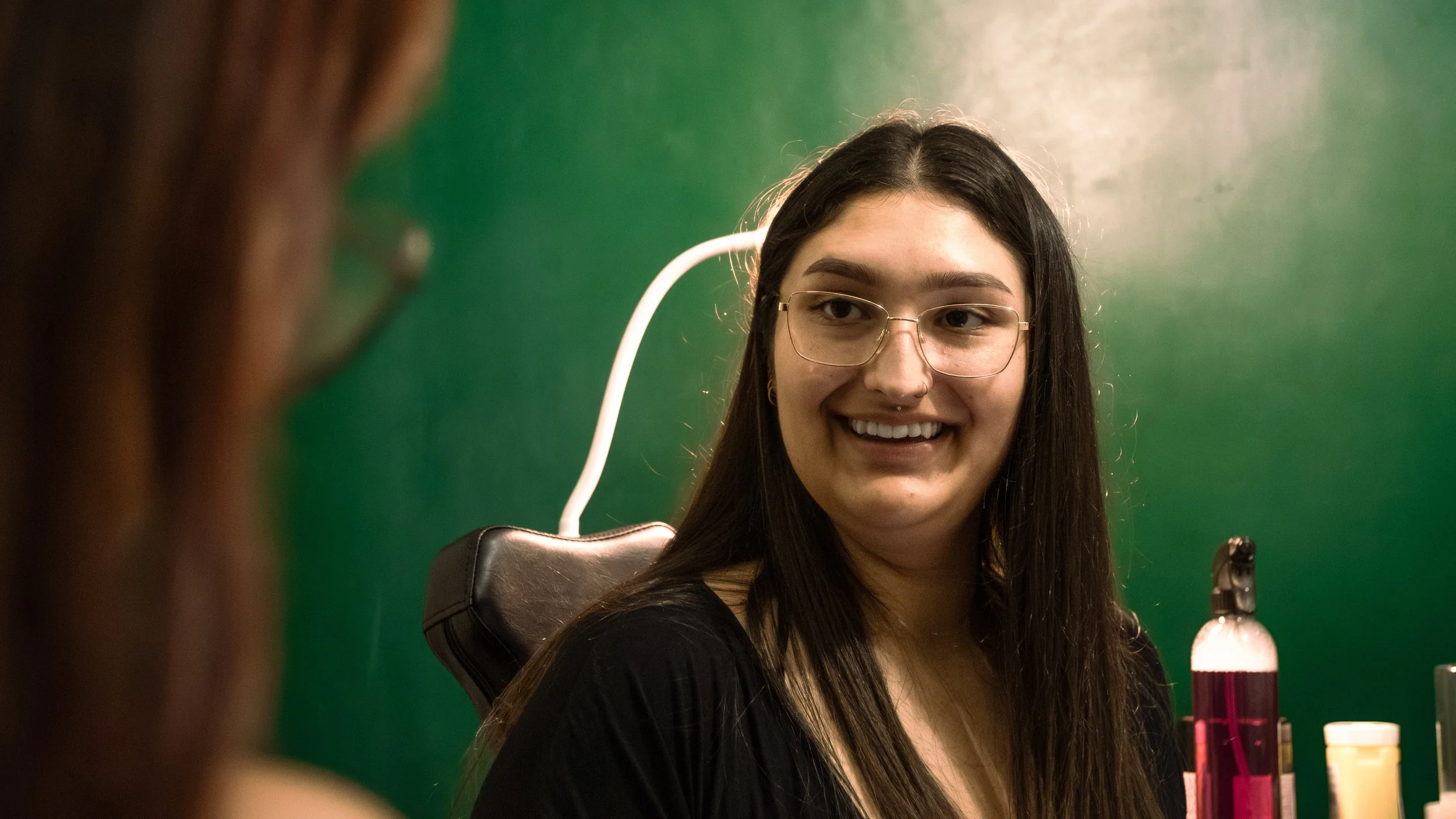 A young woman with long dark hair, glasses, and a nose piercing smiling at the camera, in front of a green background, with test tubes and bottles on a table.