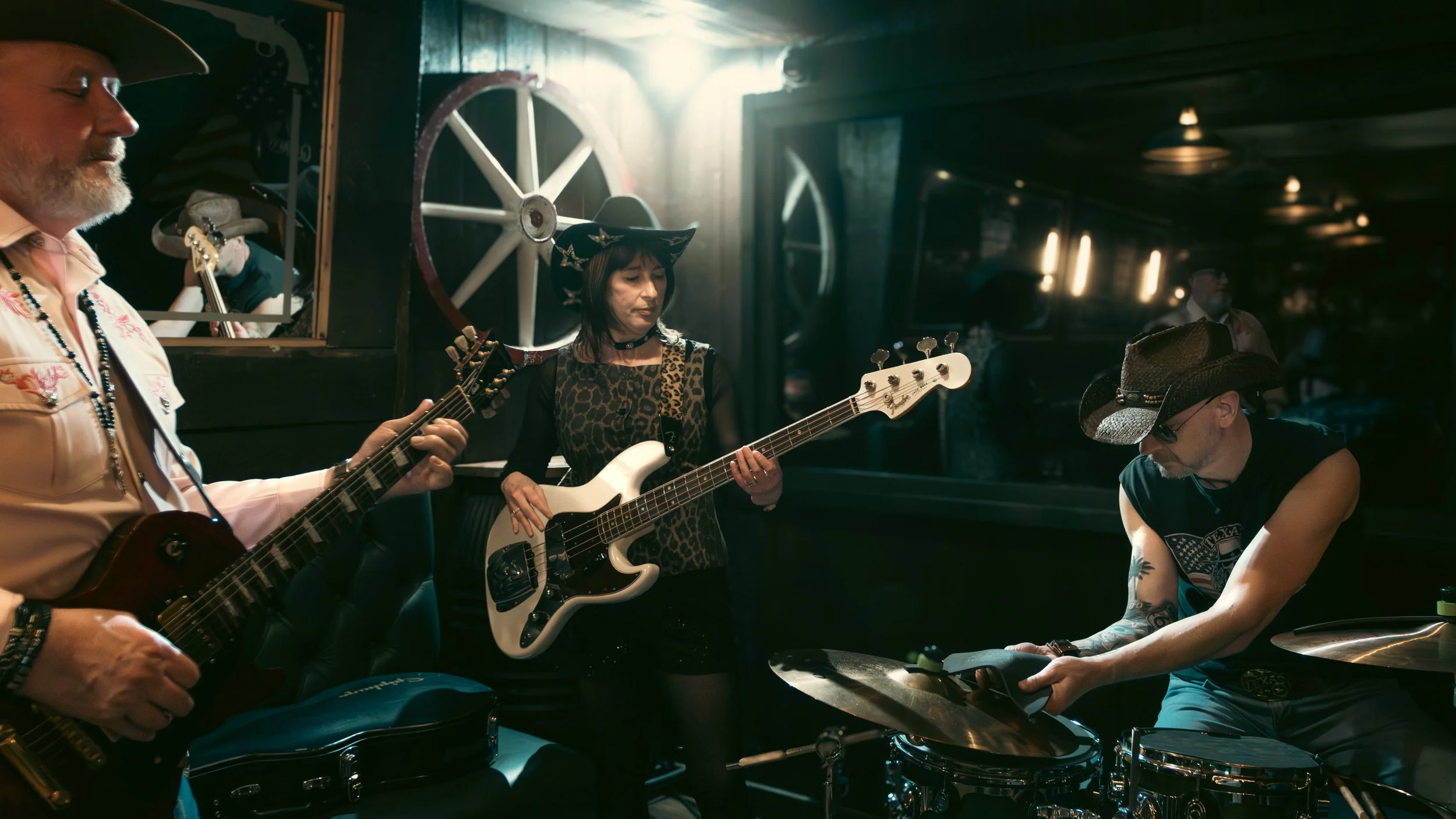 A band of four musicians performing together indoors. The man on the left is playing an electric guitar, the woman in the center is playing a bass guitar and wearing a witch hat and leopard print shirt, the man on the right is wearing sunglasses and 
