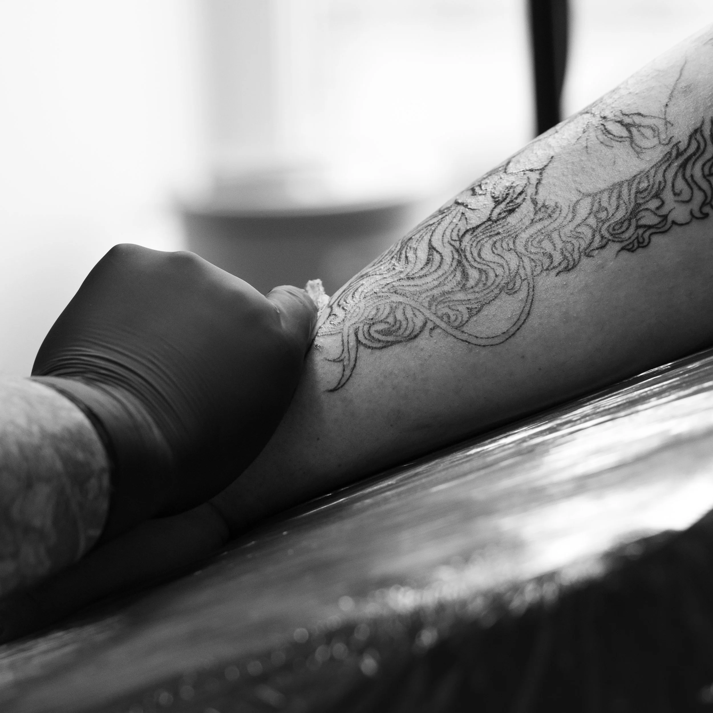 Close-up of a tattoo on a person's arm with detailed black ink line work and shading, with a blurred background.