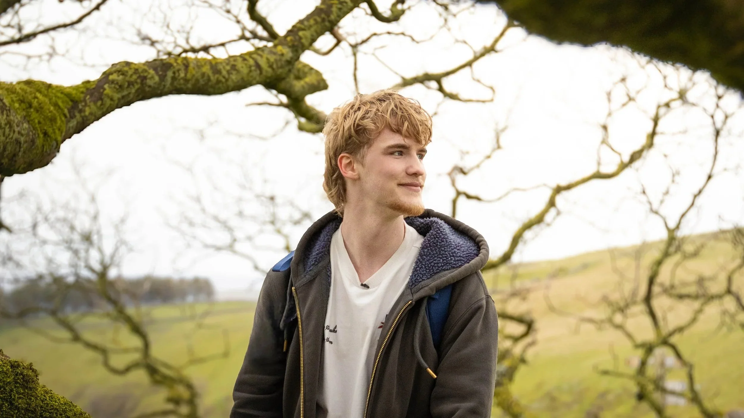 A young man with curly red hair and a slight beard standing outdoors near leafless trees, wearing a dark jacket with a hood, a white T-shirt, and a blue backpack, with a grassy field in the background.