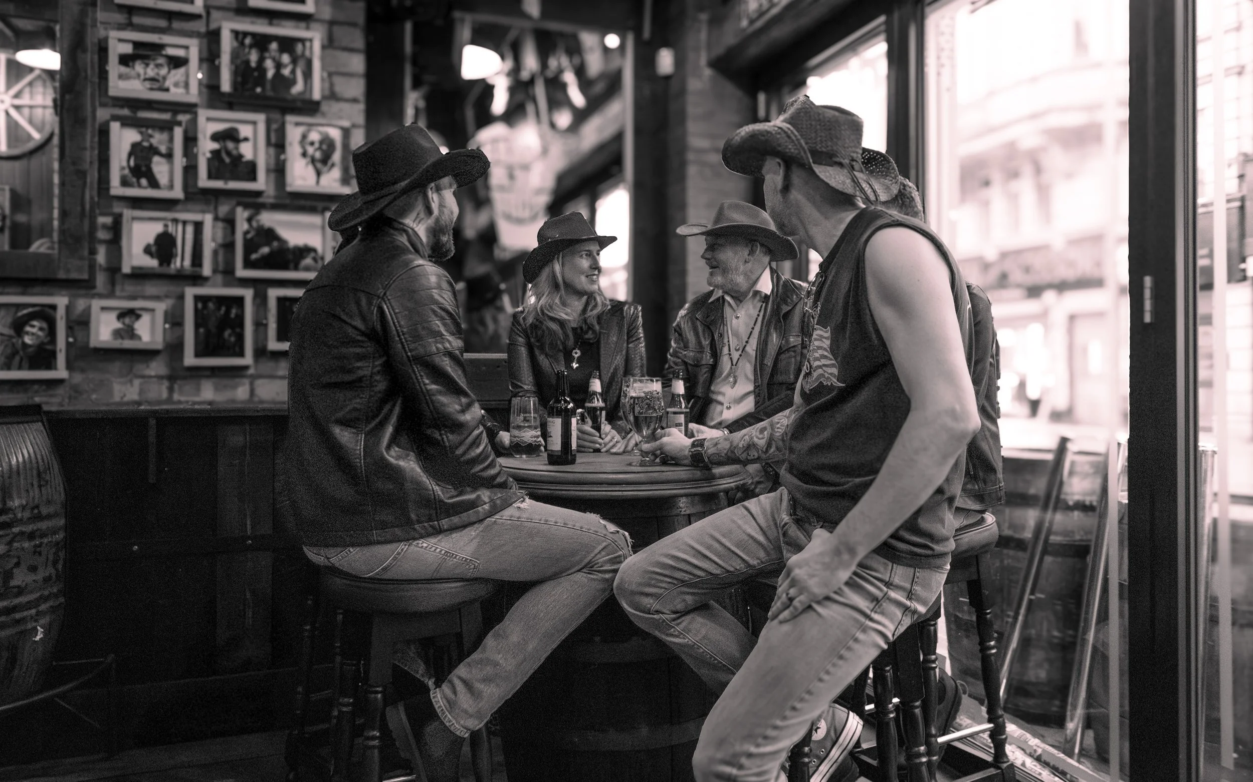 Five friends wearing cowboy hats and casual clothes sitting and standing around a table with drinks in a rustic bar, with a wall decorated with framed artwork in the background.