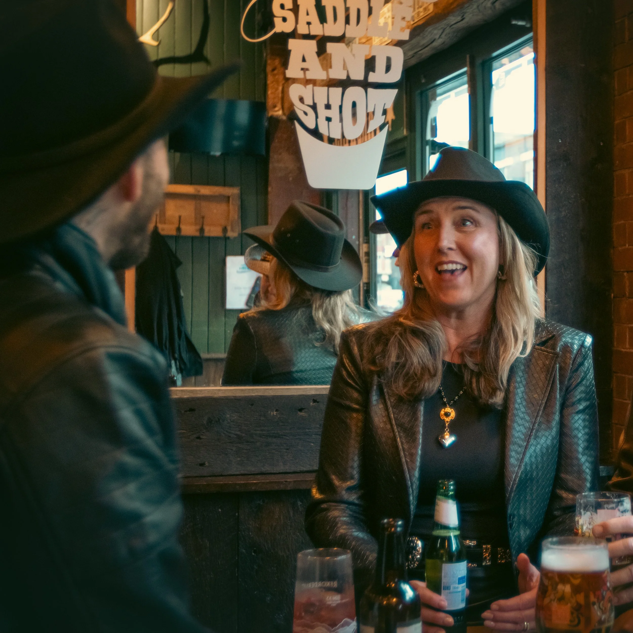 People celebrating at a bar, wearing cowboy hats, with a sign that says 'SaturdayAndShot' in the background, holding drinks.