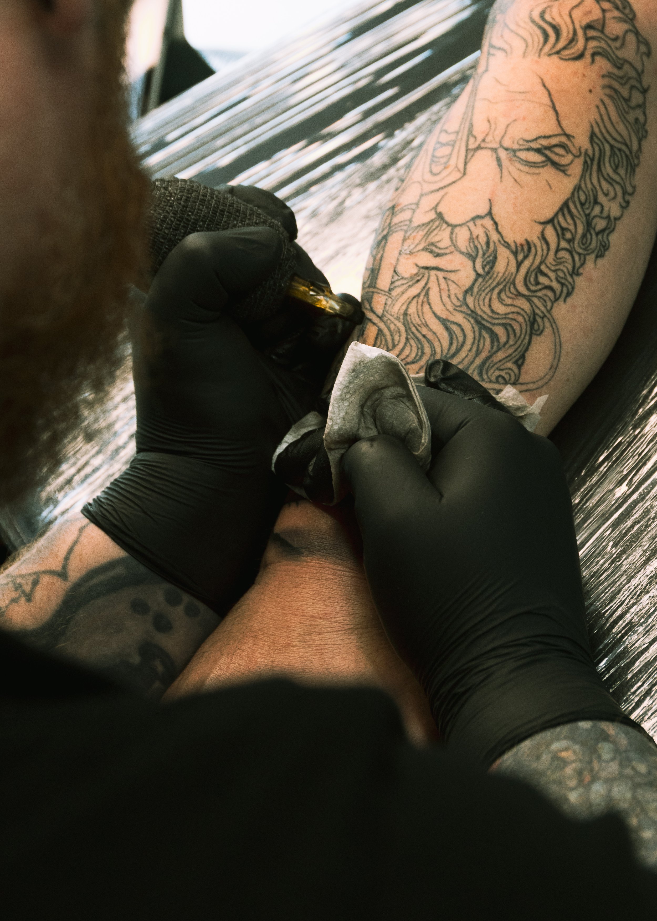 Person getting a tattoo of a detailed face with a beard and glasses on their arm, with tattoo artist wearing black gloves working on the tattoo.