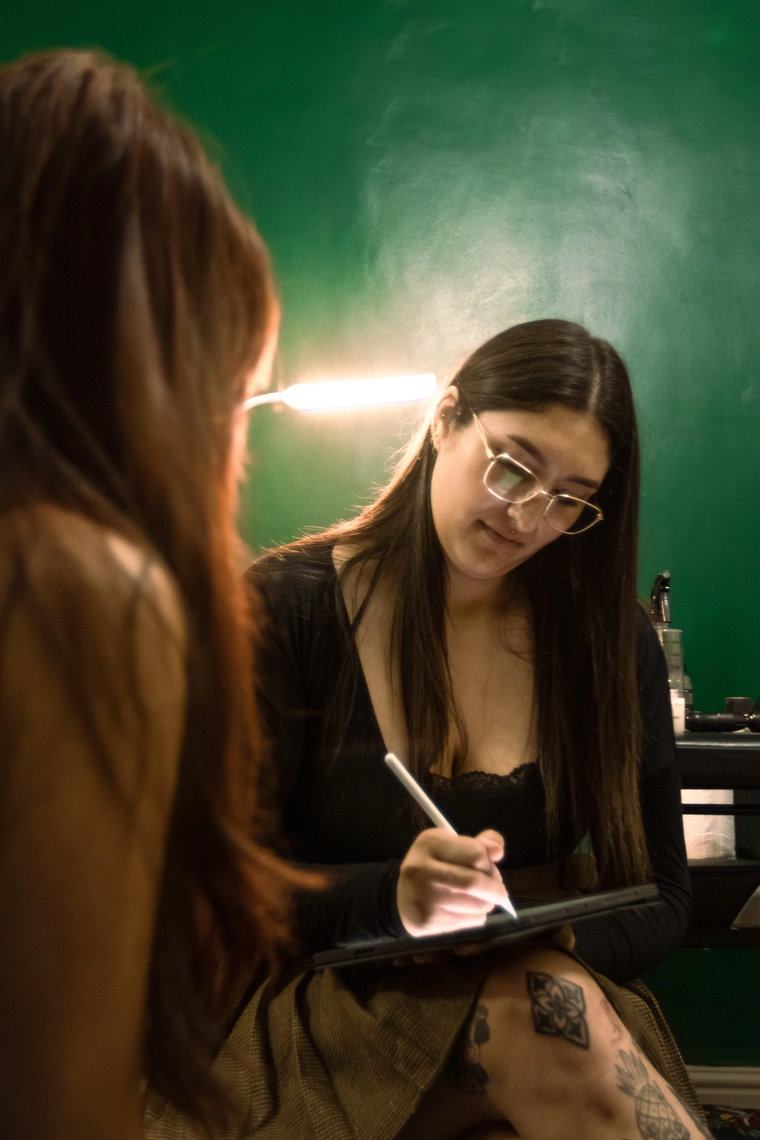 Two young women sitting at a counter, one with glasses and long dark hair, writing or drawing on a notebook, and another with reddish hair and tattoos on her leg, with a green wall background and a bright light overhead.