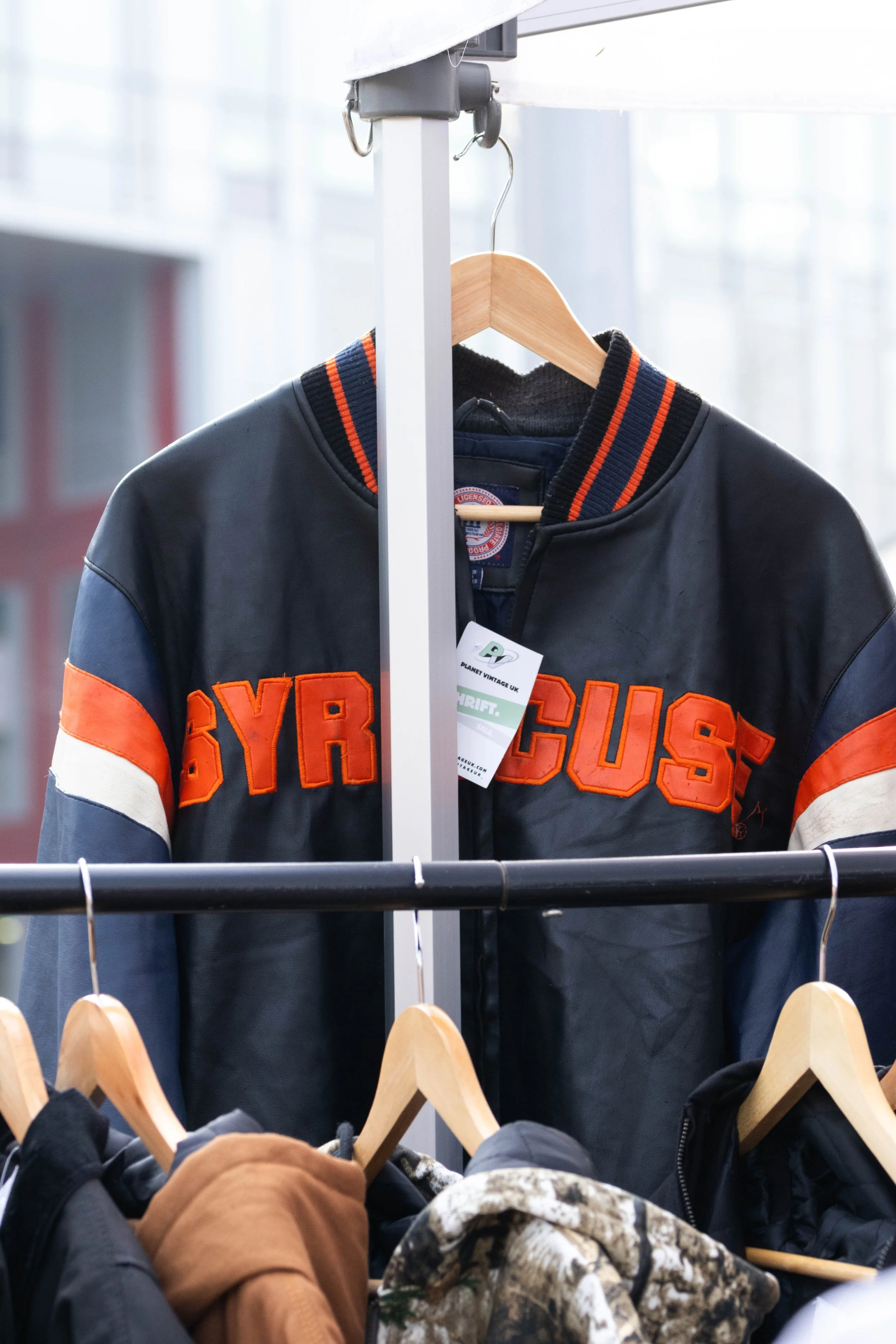Black and orange Syracuse varsity jacket hanging on a wooden hanger in a retail store with other jackets on hangers below.