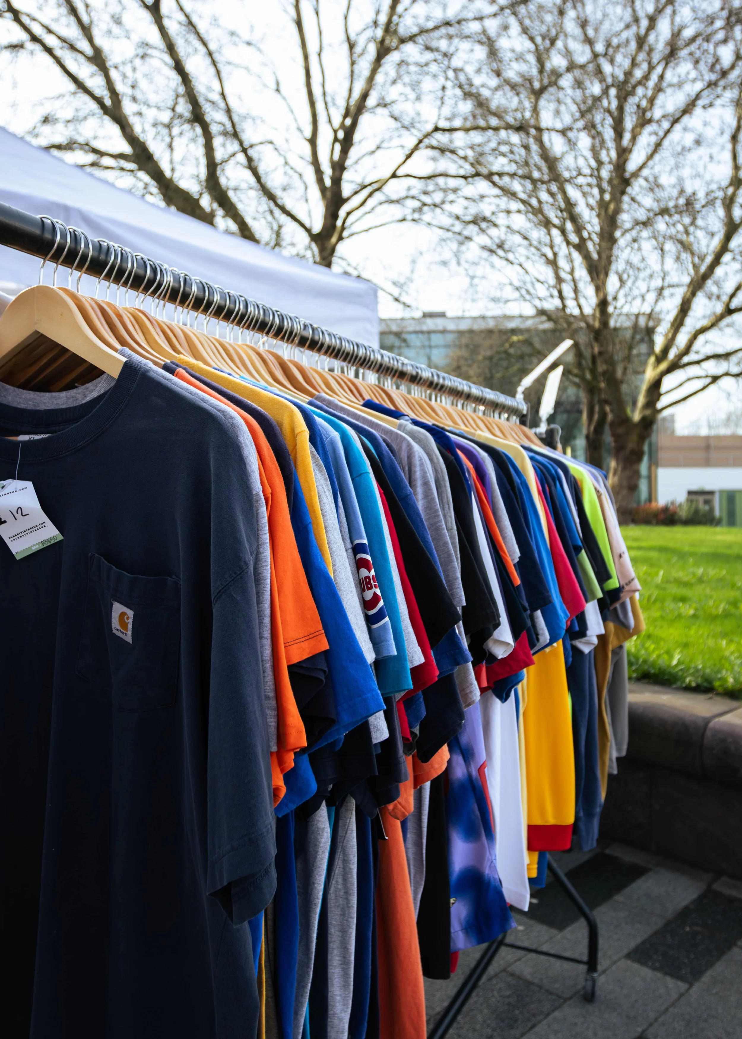 Clothing rack outdoors with multicolored T-shirts on hangers and leafless trees in the background.