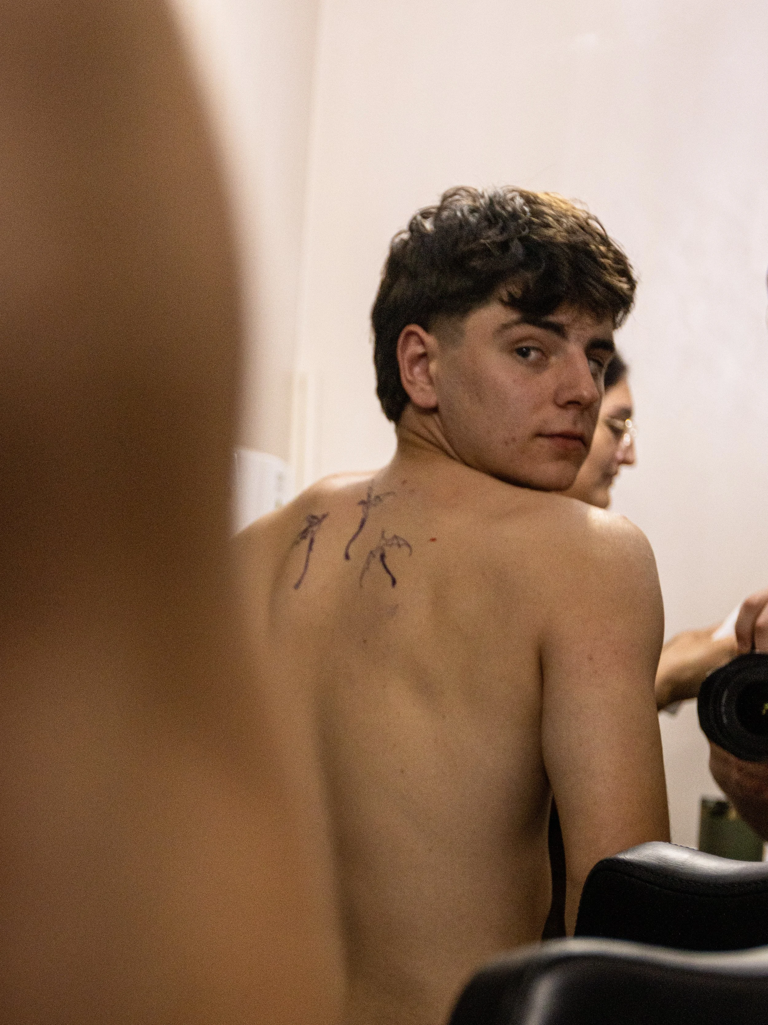 A shirtless young man with short curly hair looking over his shoulder at the camera, with ink markings on his back, in a room with beige walls and black chairs.