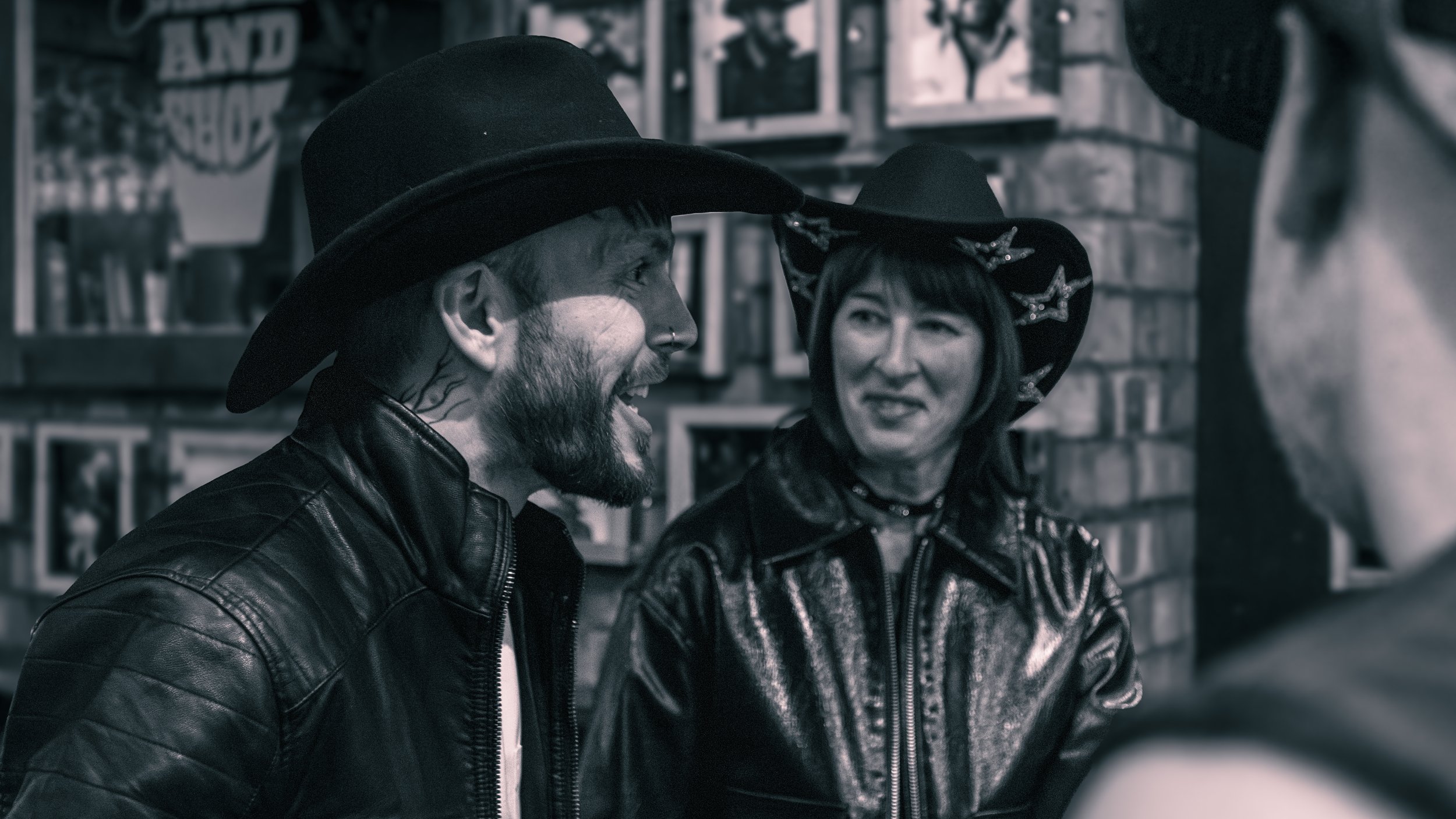 Two people wearing leather jackets and hats are in conversation in a store with picture frames on the wall.