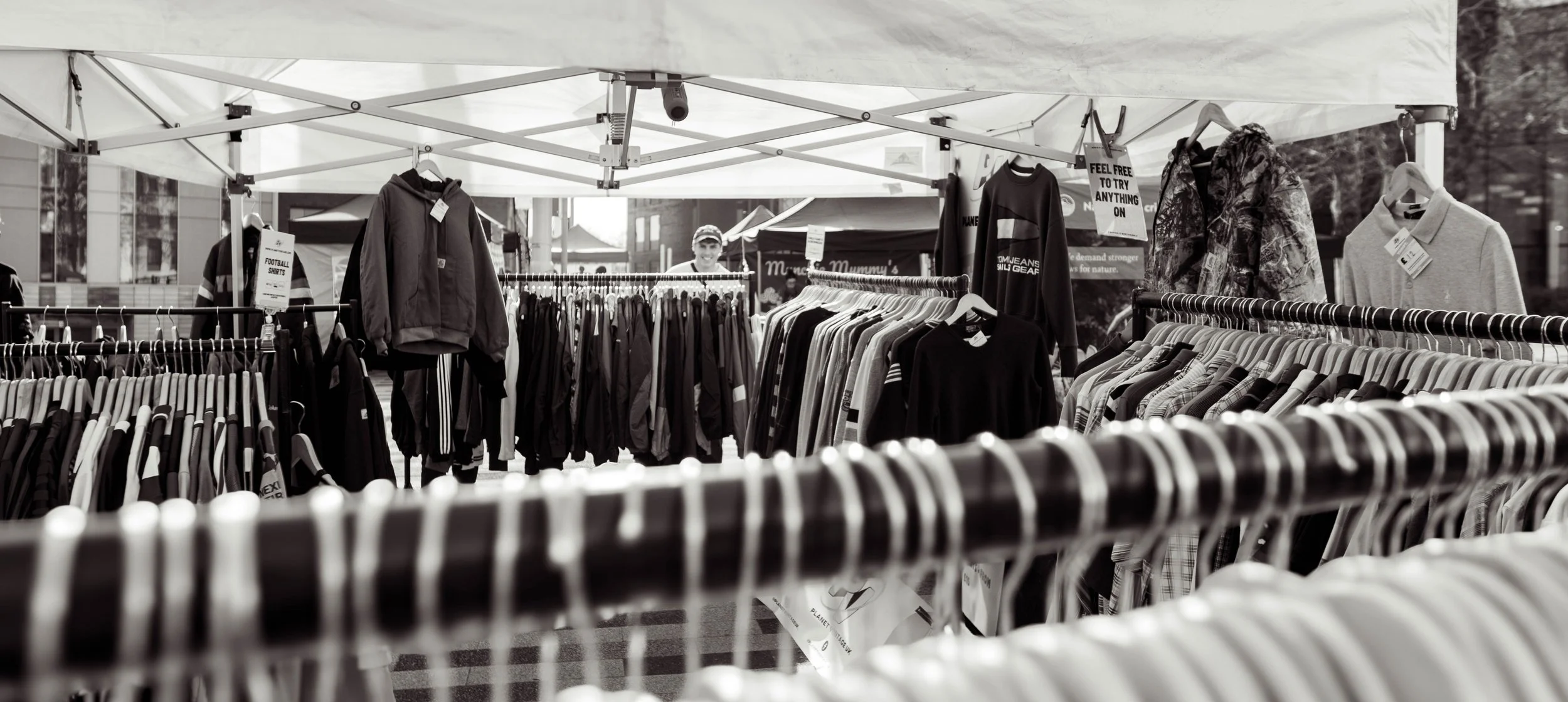 Black and white photo of an outdoor clothing market stall with various garments on racks, including jackets, hoodies, and shirts, with a person smiling in the background.