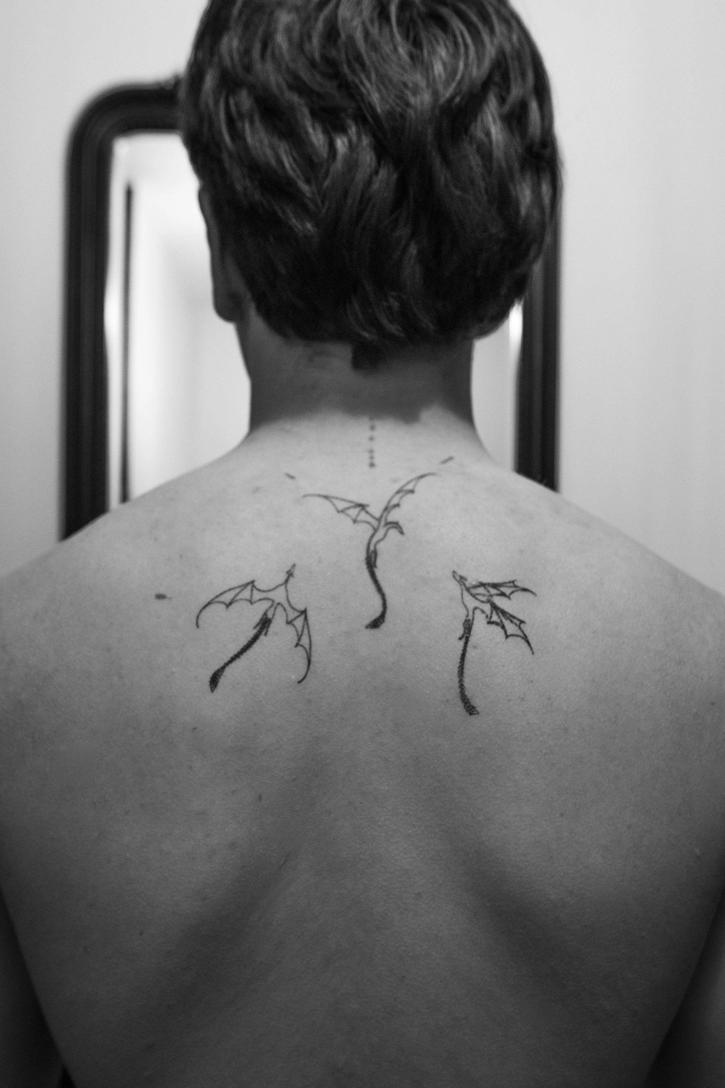 Back of a person's neck and shoulders with a tattoo of bats flying among clouds on the upper back, viewed in black and white.