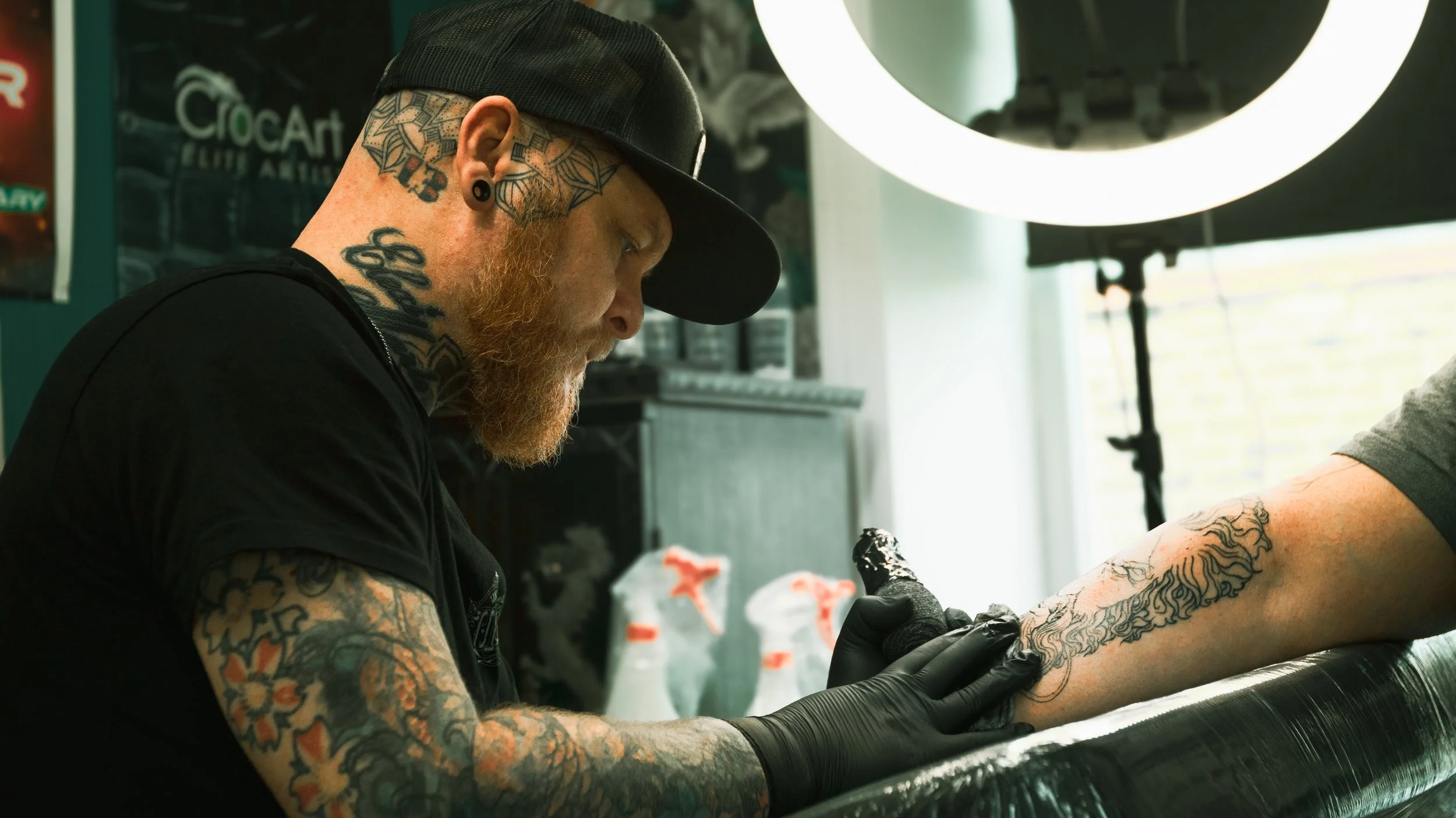 A man with tattoos and a black cap tattooing a person's arm in a tattoo studio, with bright white lighting overhead.