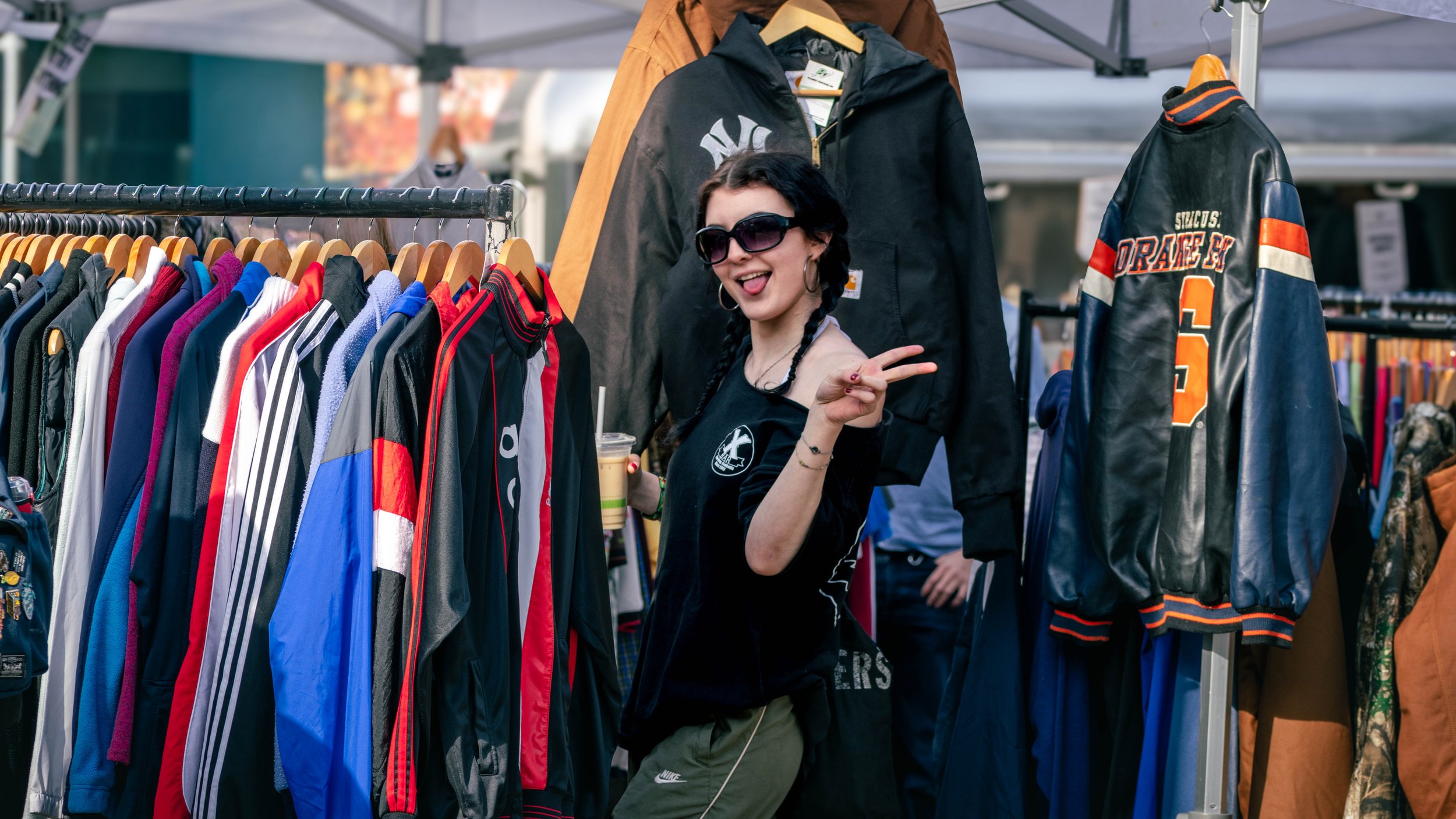 A woman with black hair, wearing sunglasses and a black shirt, shopping at an outdoor clothing market. She is smiling and making a peace sign with her hand, standing in front of racks of jackets and sweaters.