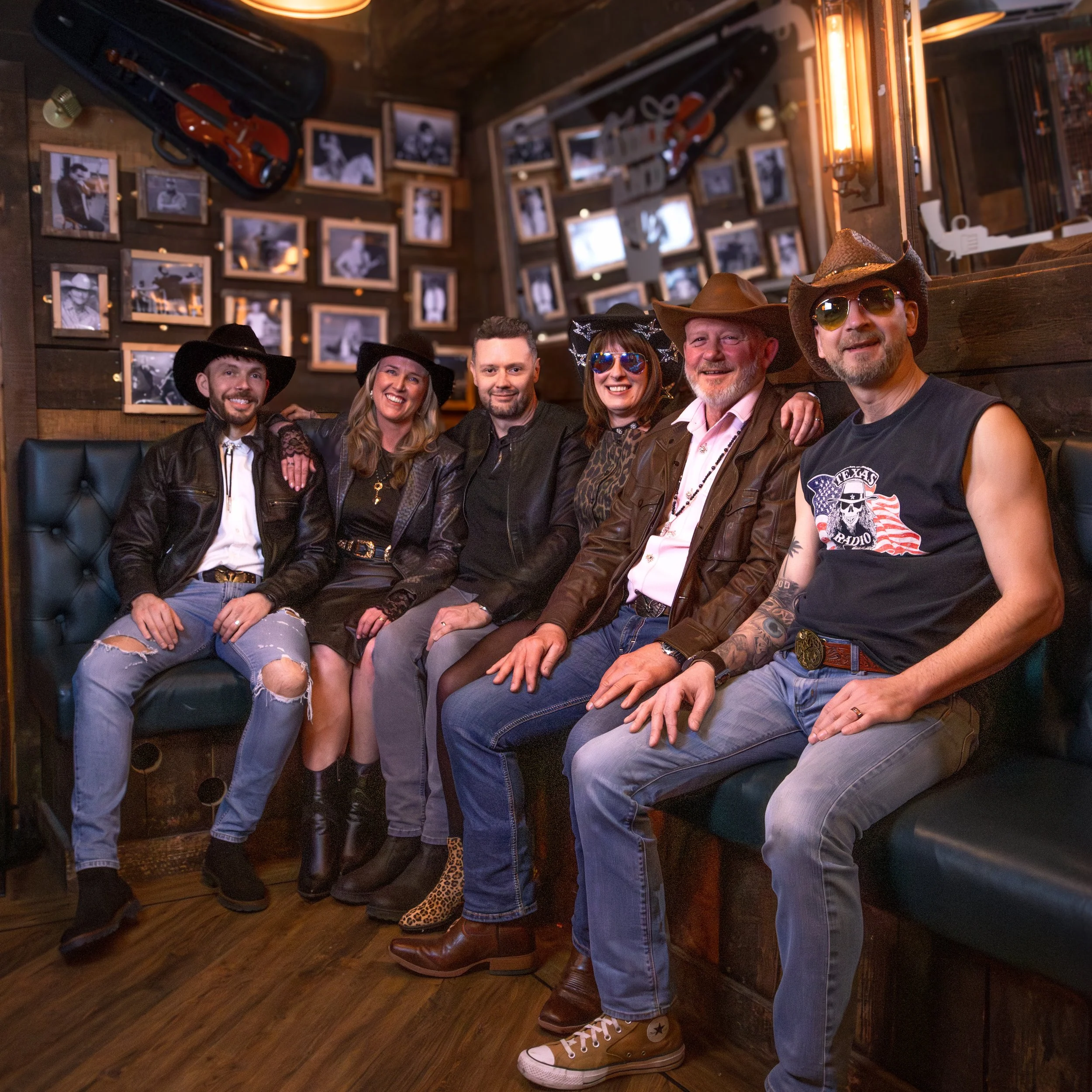 A group of six people dressed in casual cowboy attire, including hats and sunglasses, sitting close together on a bench inside a rustic bar or restaurant with wooden walls decorated with small framed photographs and guitars.