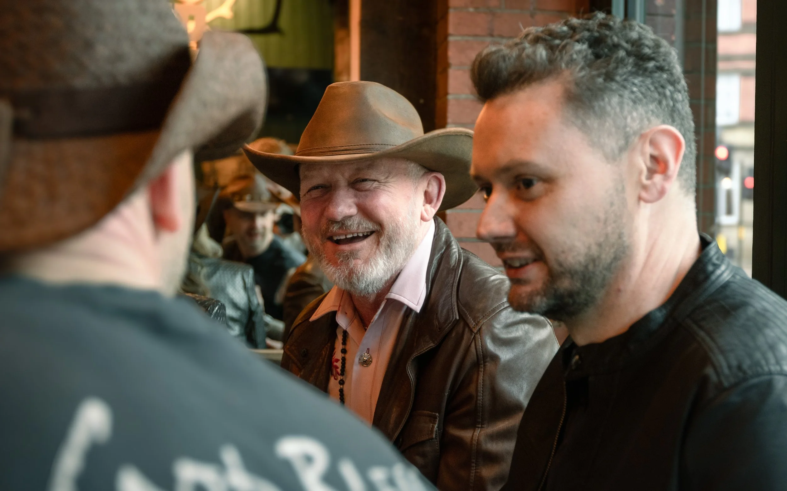 Three men sitting at a bar with smiles and engaging in conversation. One older man with a cowboy hat, a white beard, and a leather jacket. One young man with short curly hair and a black leather jacket. Another man, partially visible, wearing a dark 