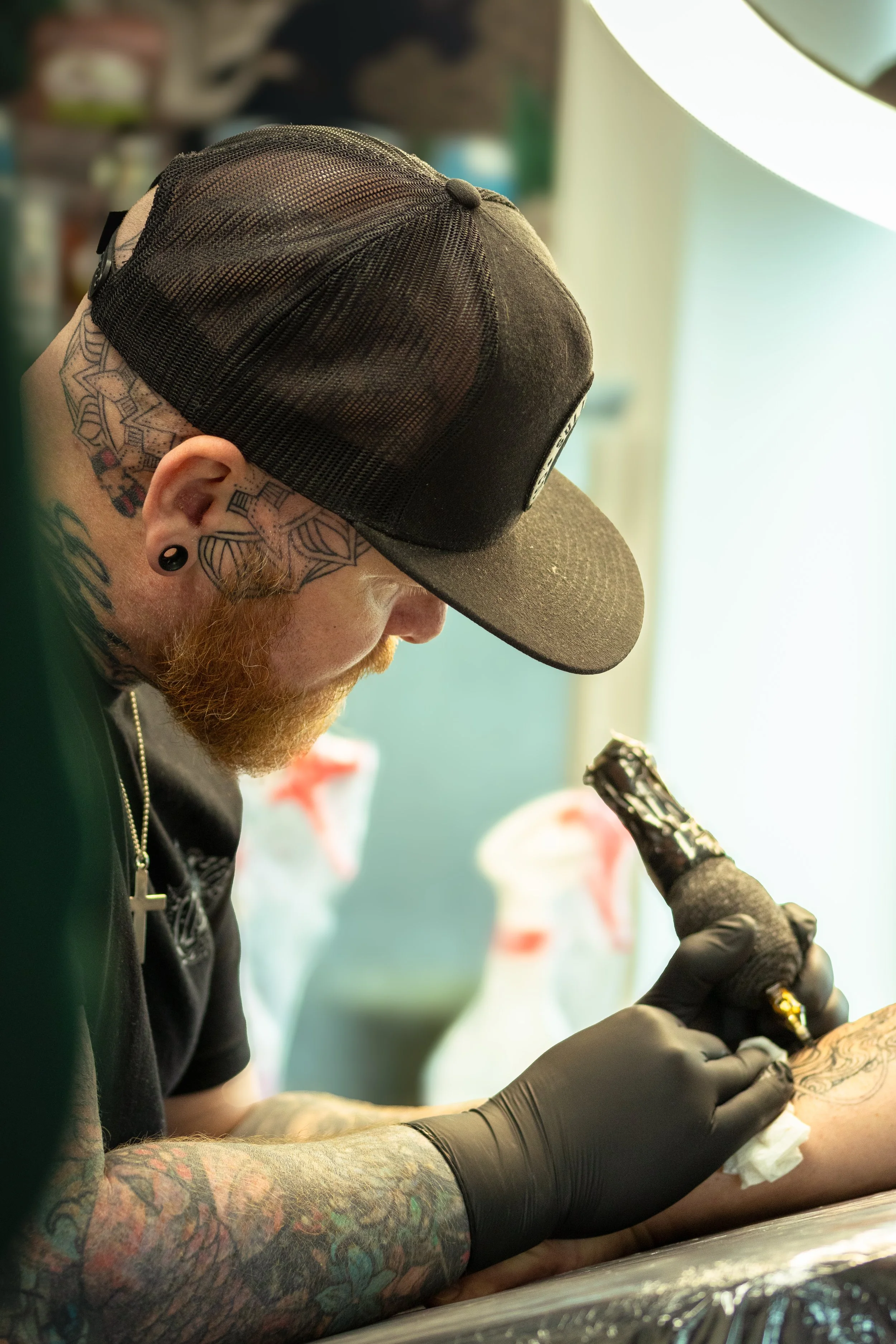 A person with tattoos on their arm and neck, wearing a black baseball cap and gloves, is tattooing or applying ink to someone's arm or leg.