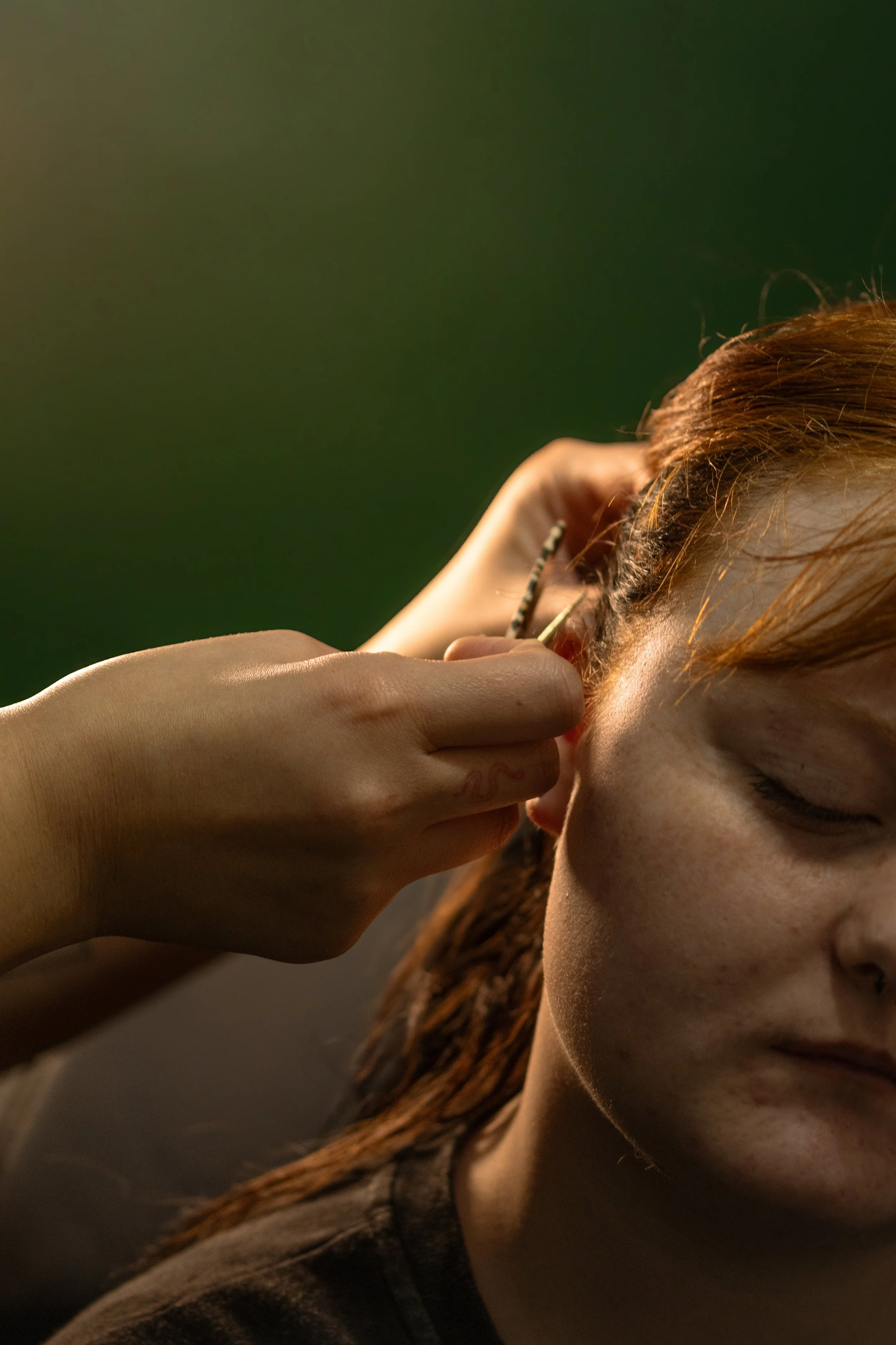 A person with red hair is inserting an earring into their ear, with a dark blurred background.