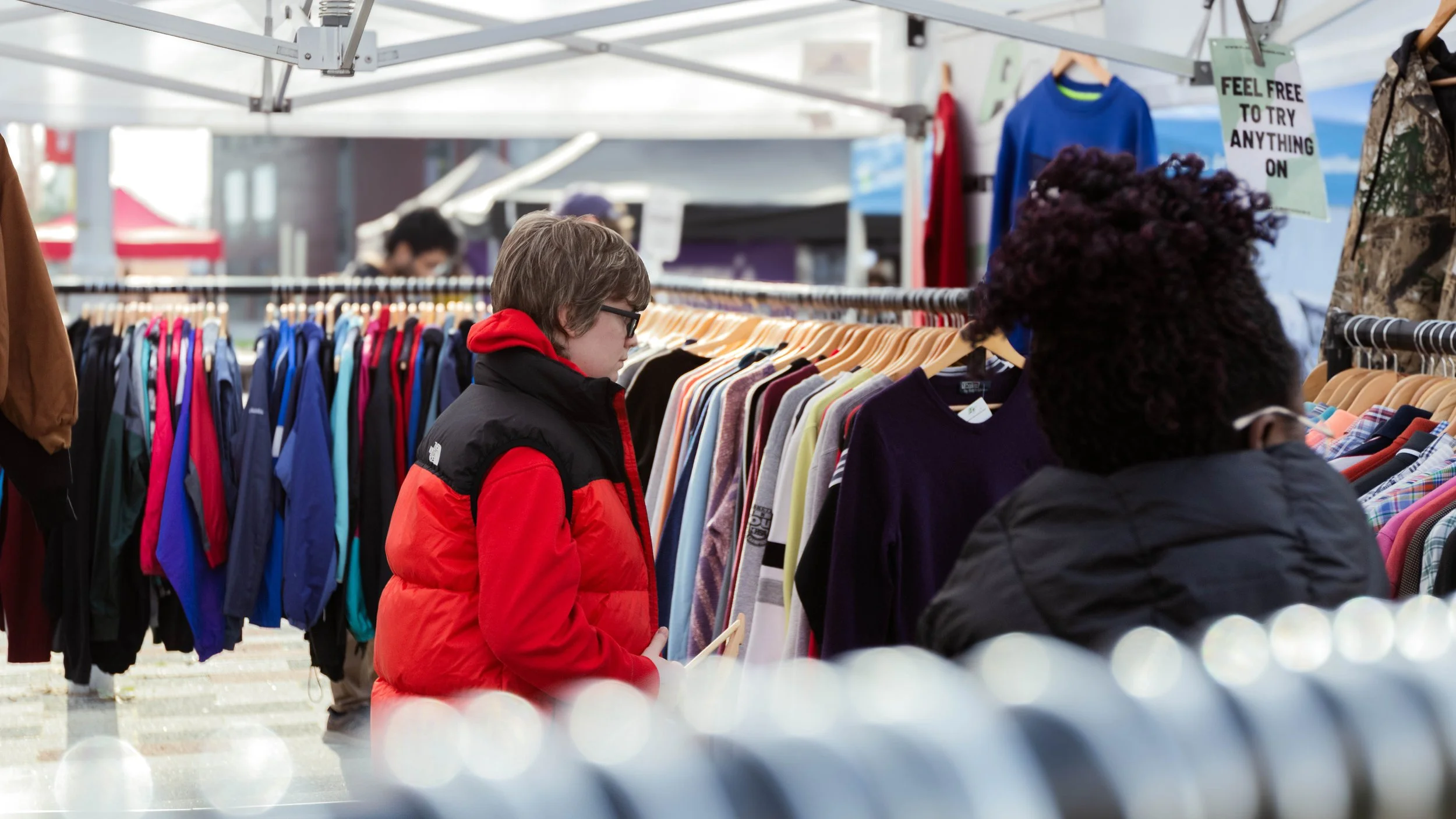People shopping for clothes at an outdoor market with racks of colorful clothing and a sign that reads 'Feel free to try anything on'.